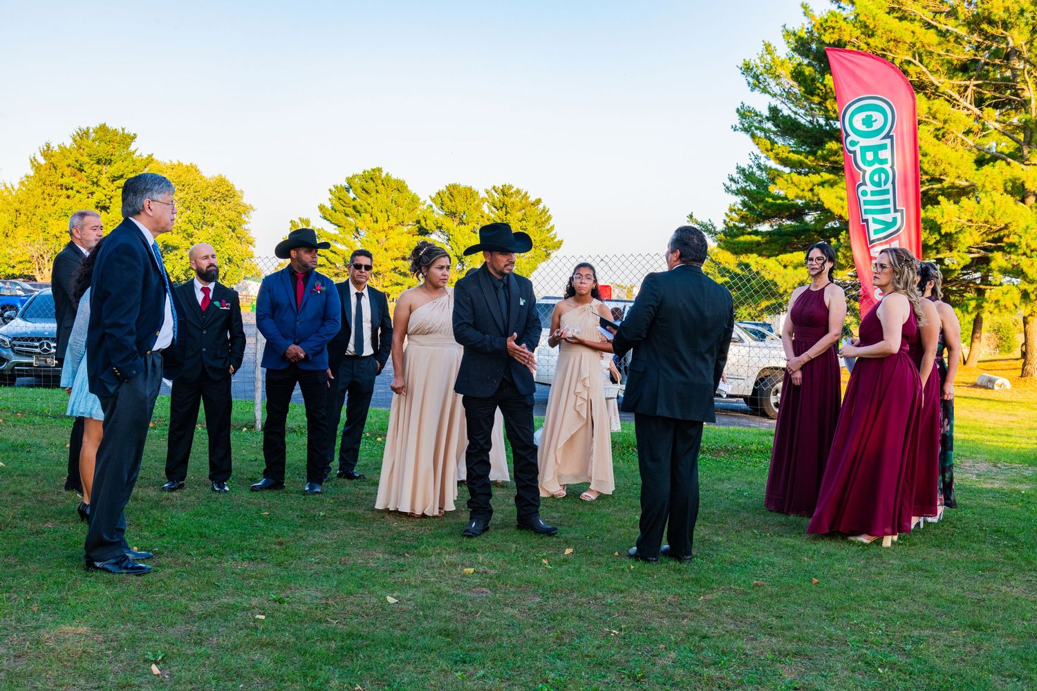 Wedding party gathered outdoors on green lawn during golden hour ceremony with colorful banner backdrop.