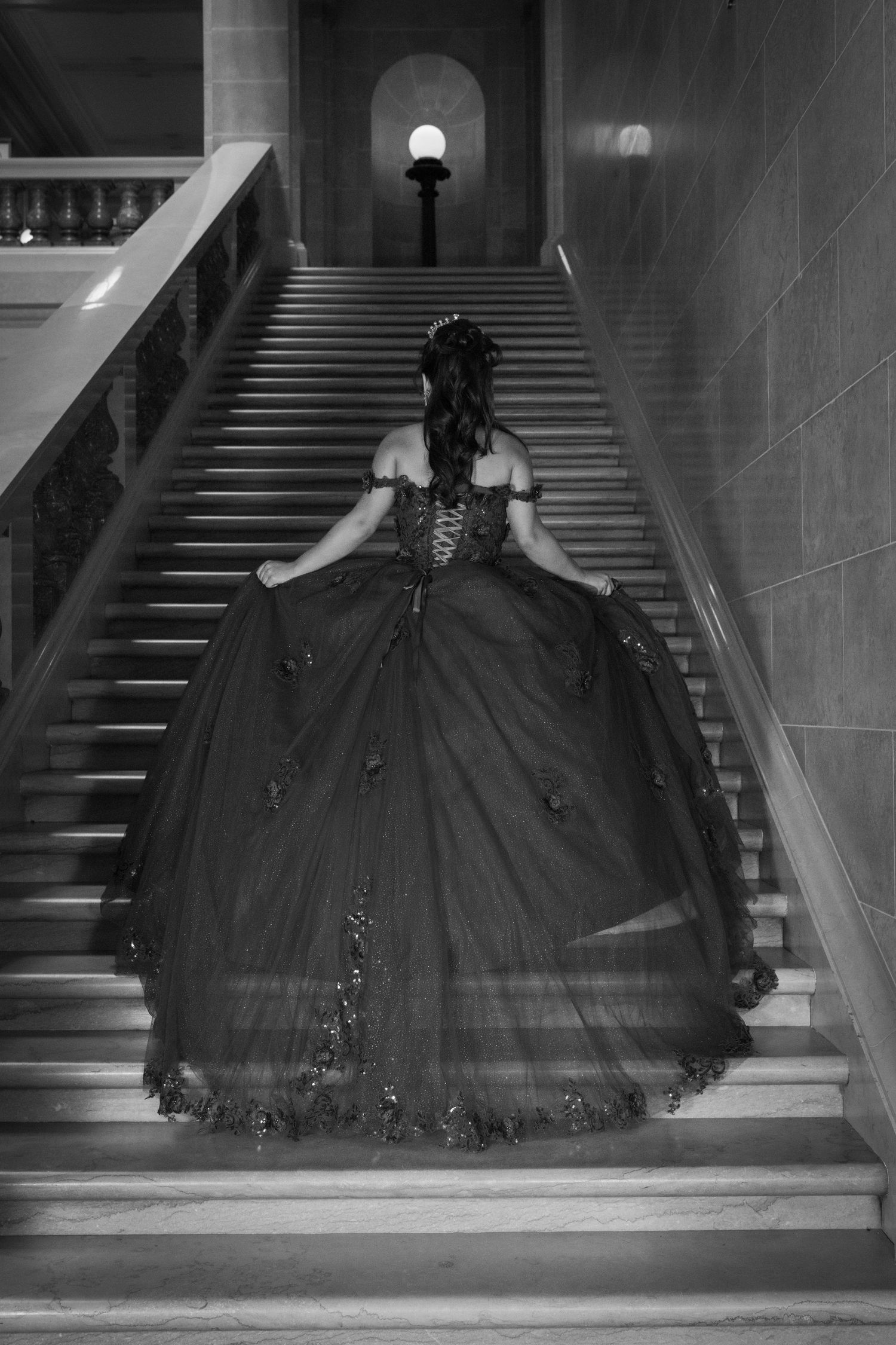 Black and white photo of woman in elaborate ball gown ascending grand staircase.