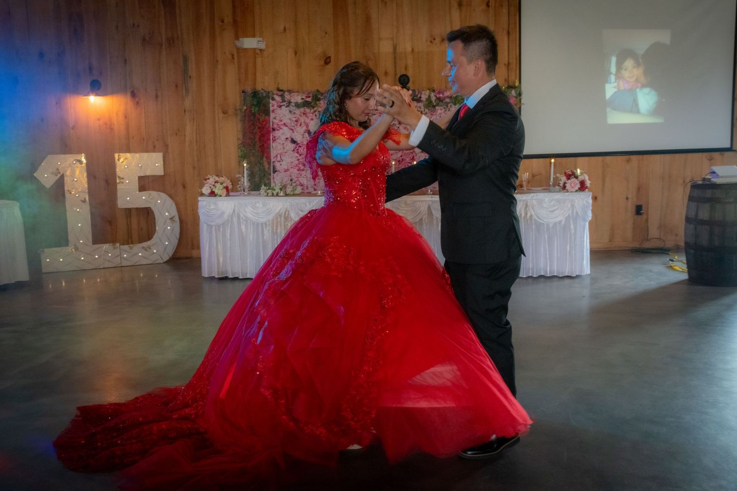 A young woman in a stunning red ball gown dances with a man at her quinceañera with a lit '15' sign.