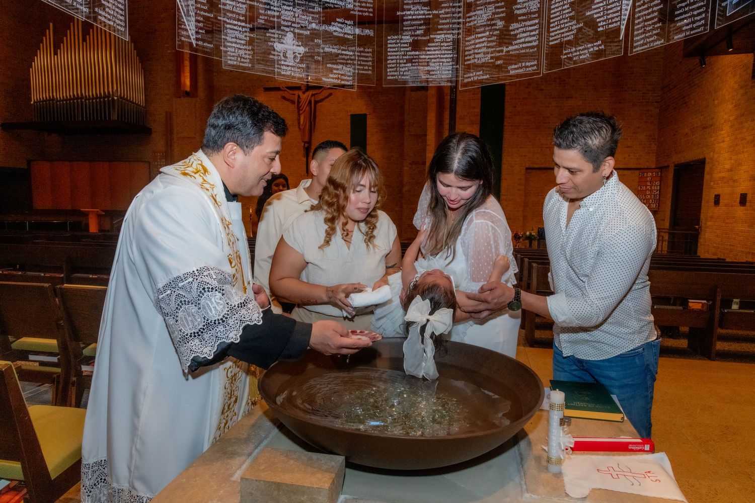 Priest performing Catholic baptism ceremony at a church font with family gathered around a baby in white.