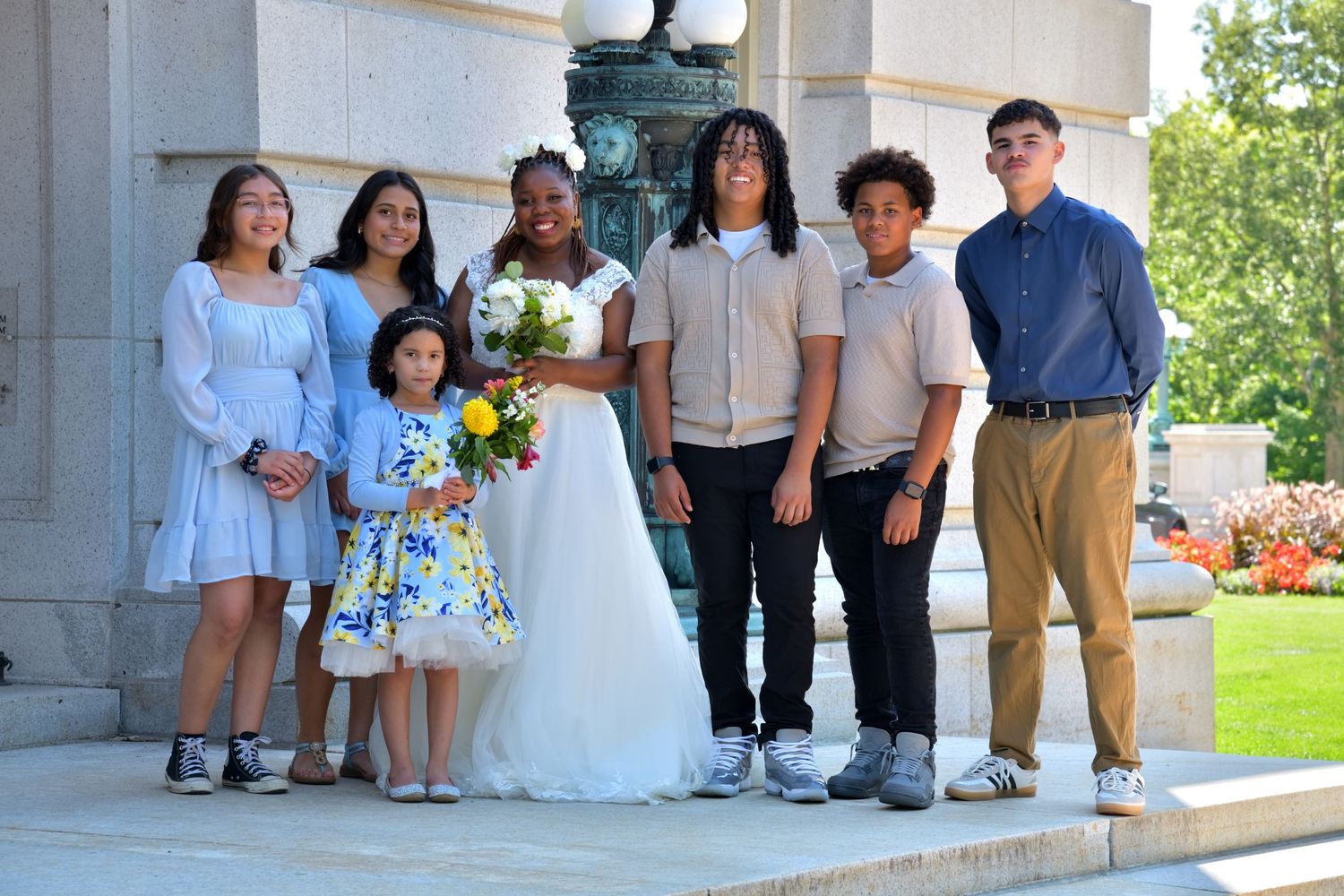 Bride in white gown poses with wedding party children outside stone building with bouquet.