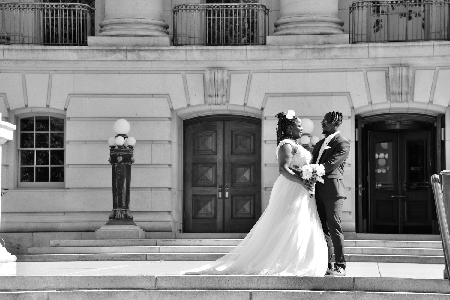 A bride and groom stand on the steps of a grand neoclassical building in a romantic black and white photo.