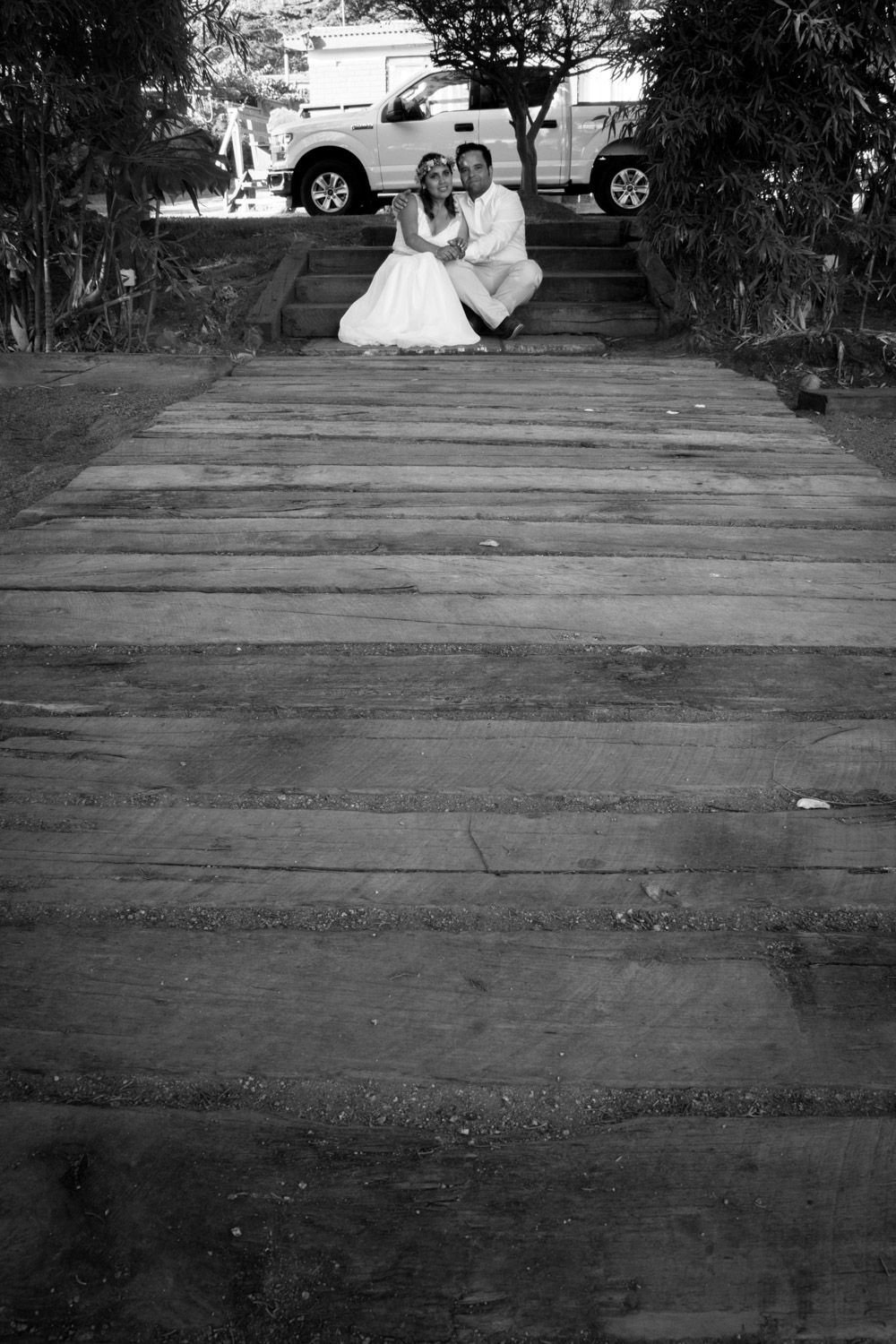 Black and white photo of bride and groom sitting on wooden steps leading to trees.