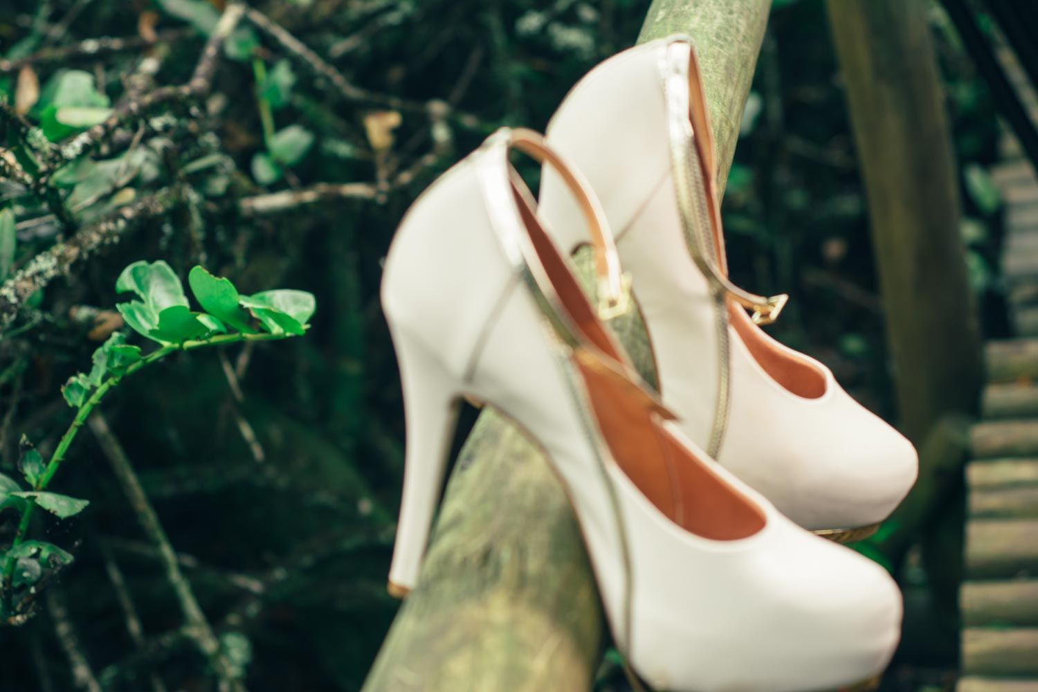 Elegant white high heel bridal shoes with gold ankle strap resting on wooden railing.