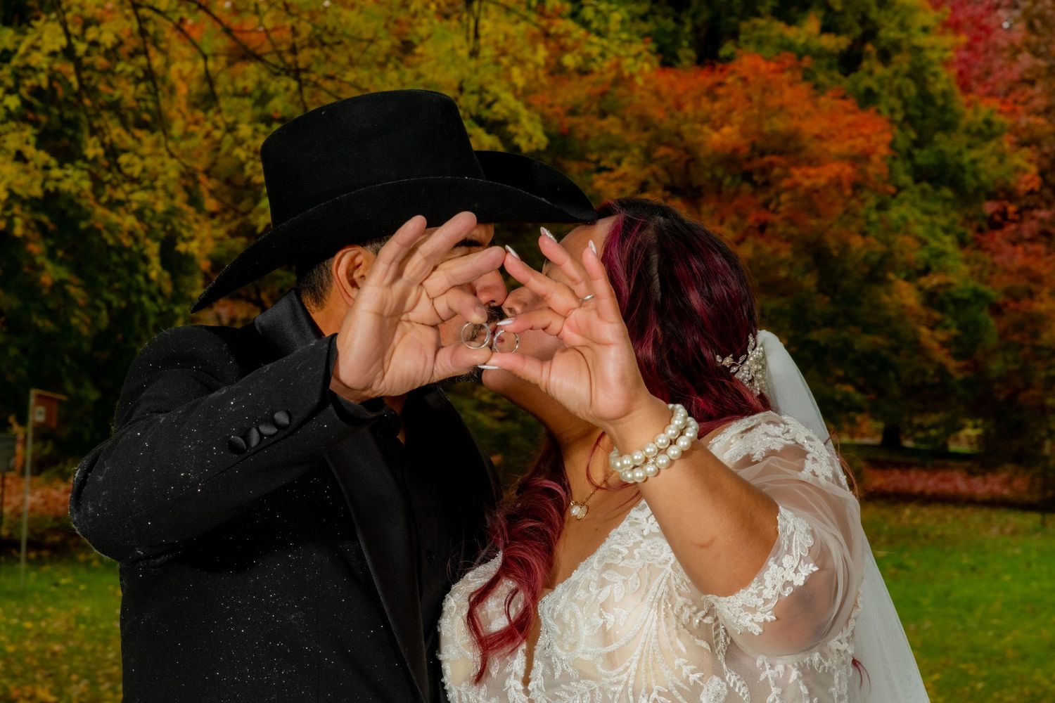 Newlyweds make heart shape with hands in front of colorful autumn trees during wedding photos.
