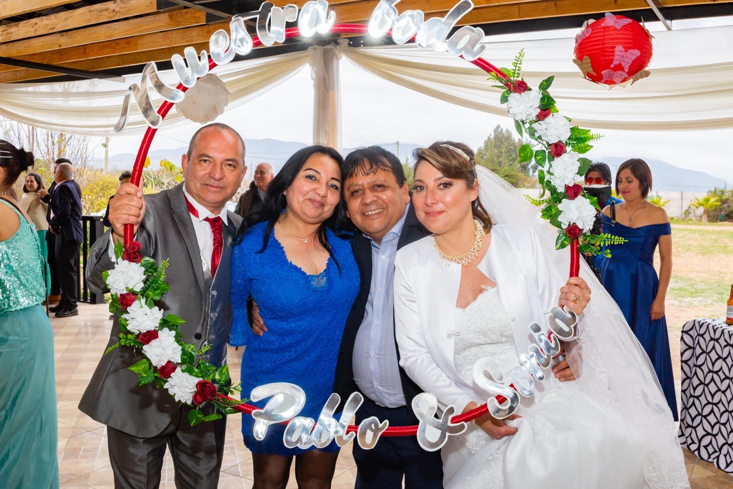 Wedding guests pose with bride and groom under decorative floral arch at outdoor ceremony.