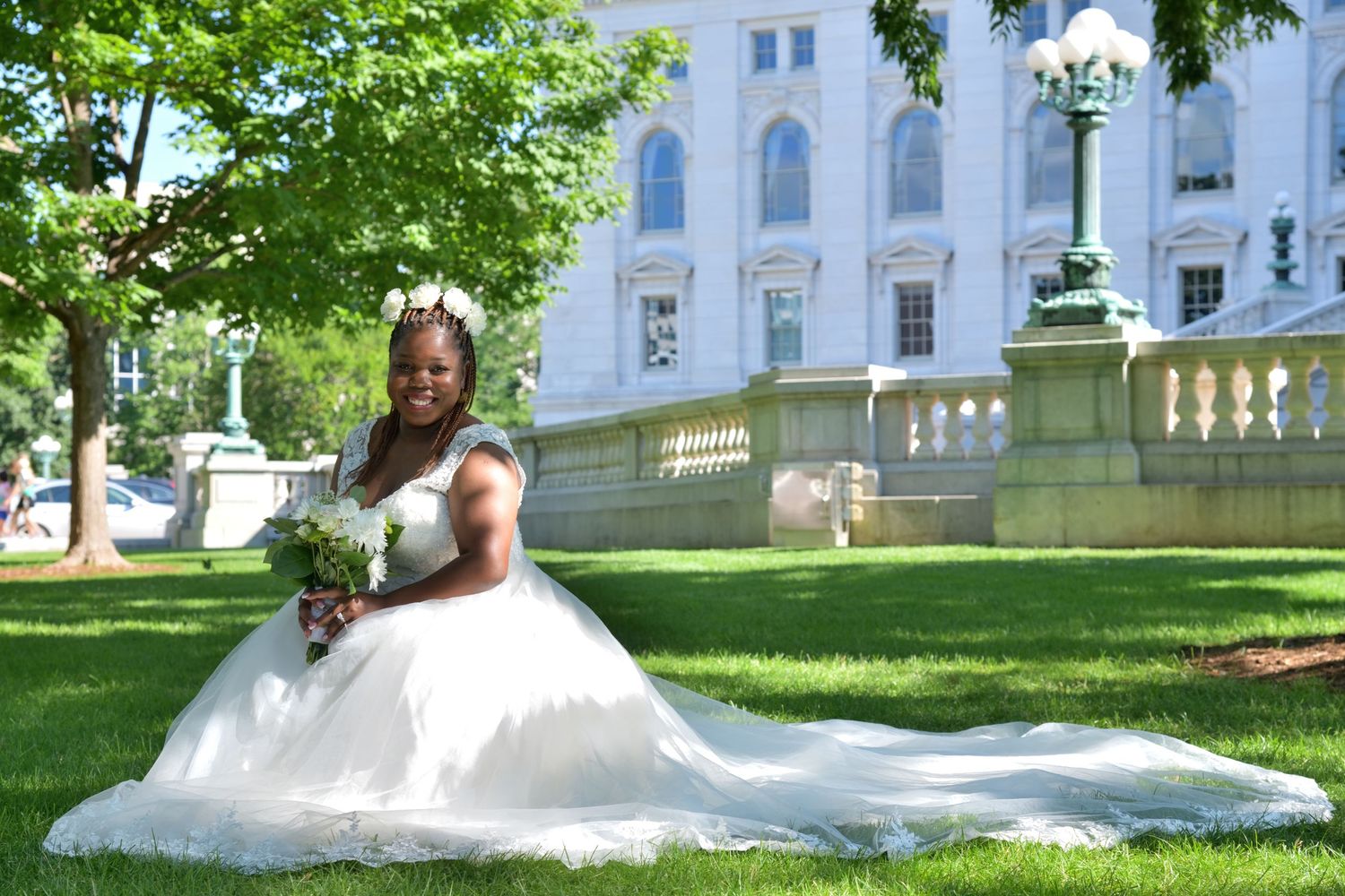 A Black bride in a flowing white ball gown sits gracefully on green grass outside a white government building.