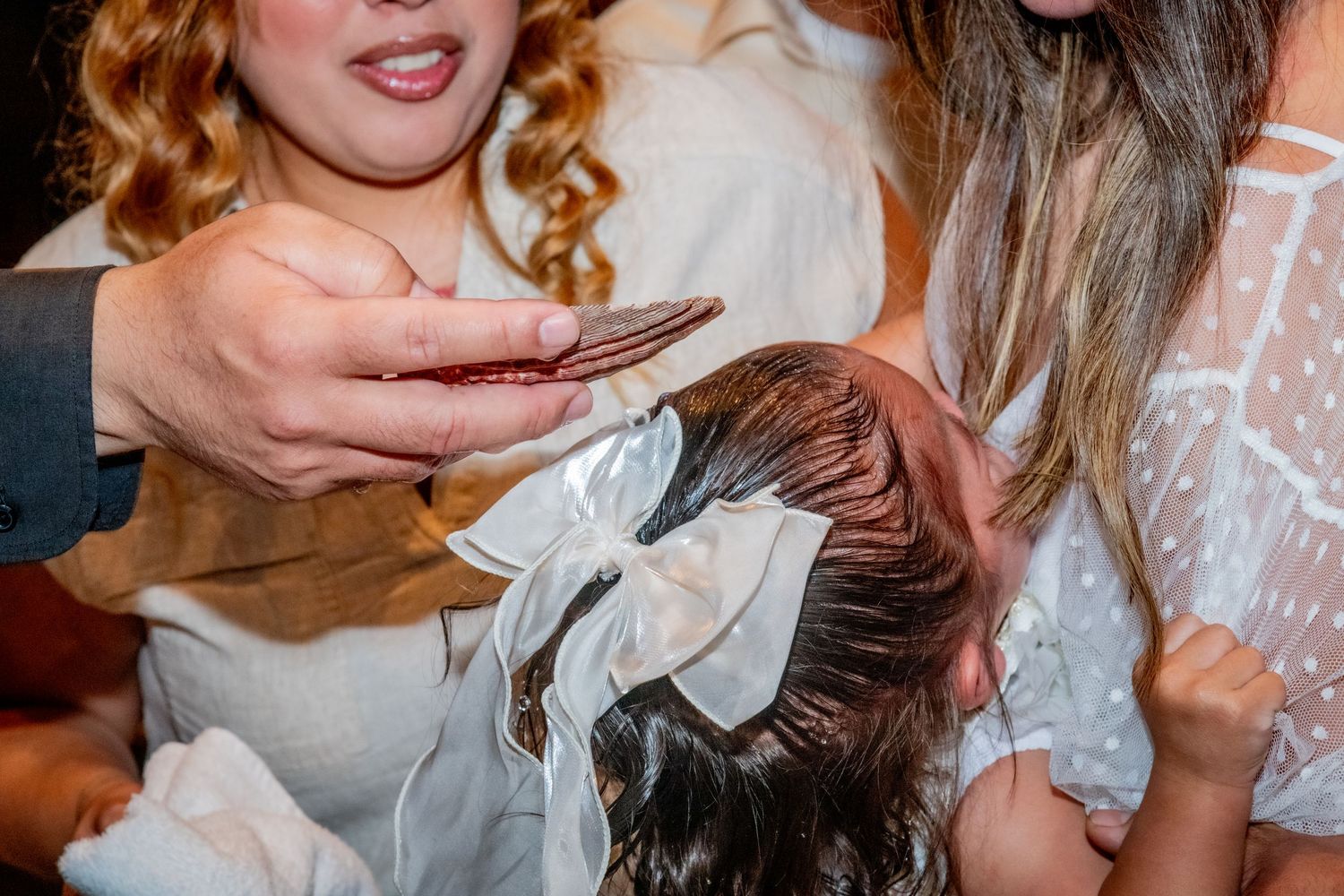 Woman with long nails styling a young girl's dark hair with a white bow at a party.