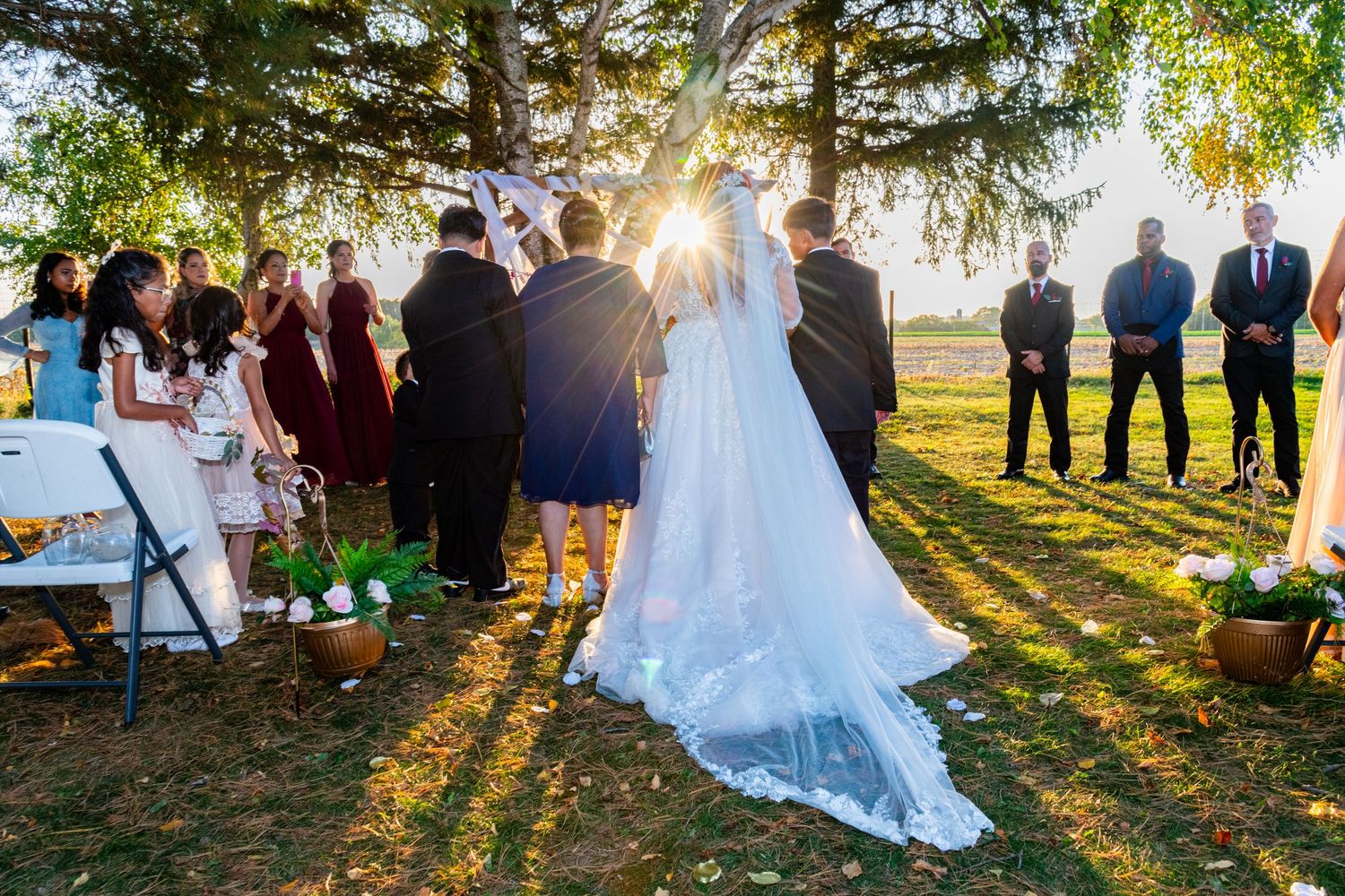 Outdoor wedding ceremony under large tree with bride in flowing veil during stunning golden sunset.
