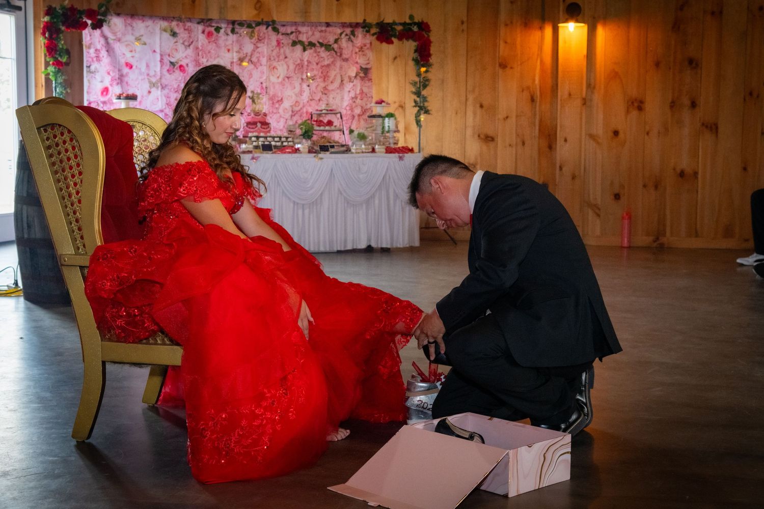 A man kneels to place a shoe on a quinceañera girl in a red gown during the shoe ceremony.