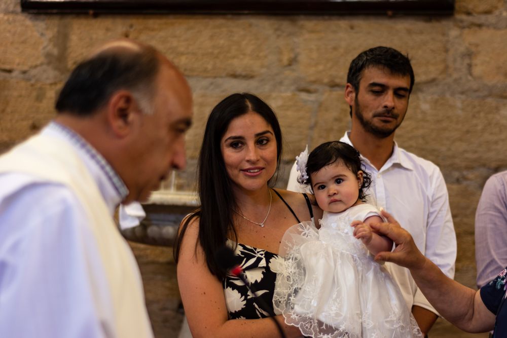 Woman in floral dress holds baby in white christening gown at a Catholic baptism ceremony.