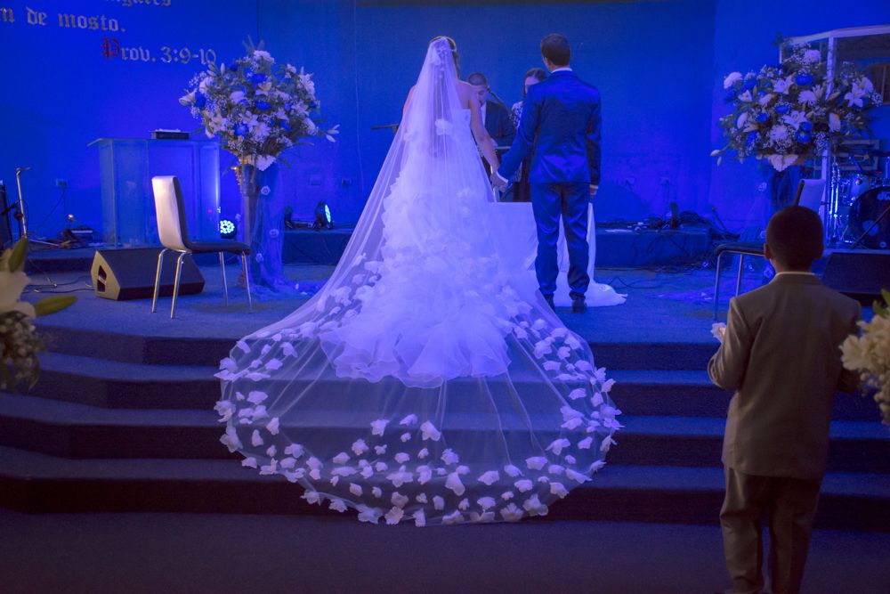 Bride in stunning floral cathedral veil stands at church altar during wedding ceremony with blue lighting.