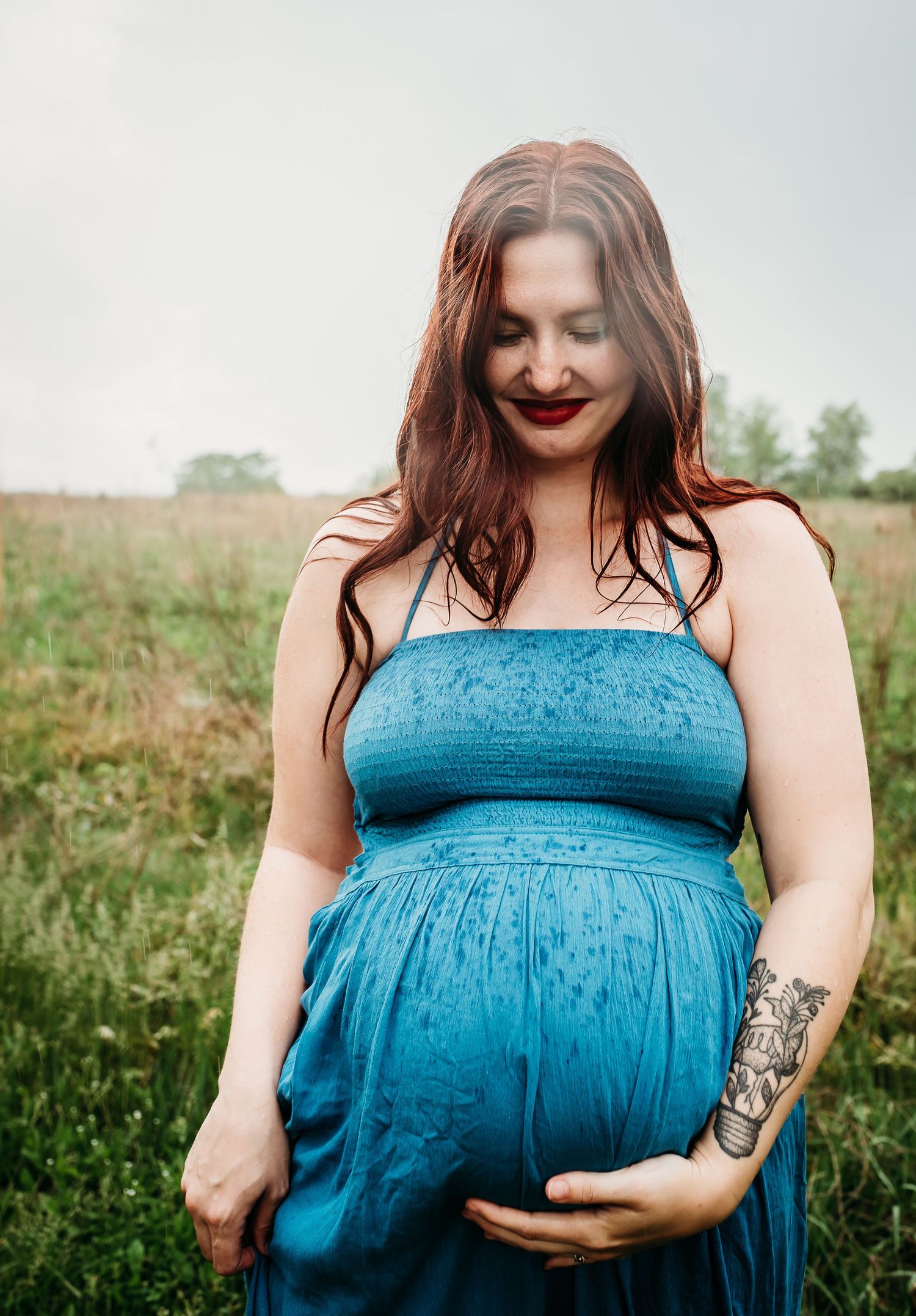 An expectant mother in a blue dress smiles while standing in a sunlit meadow.