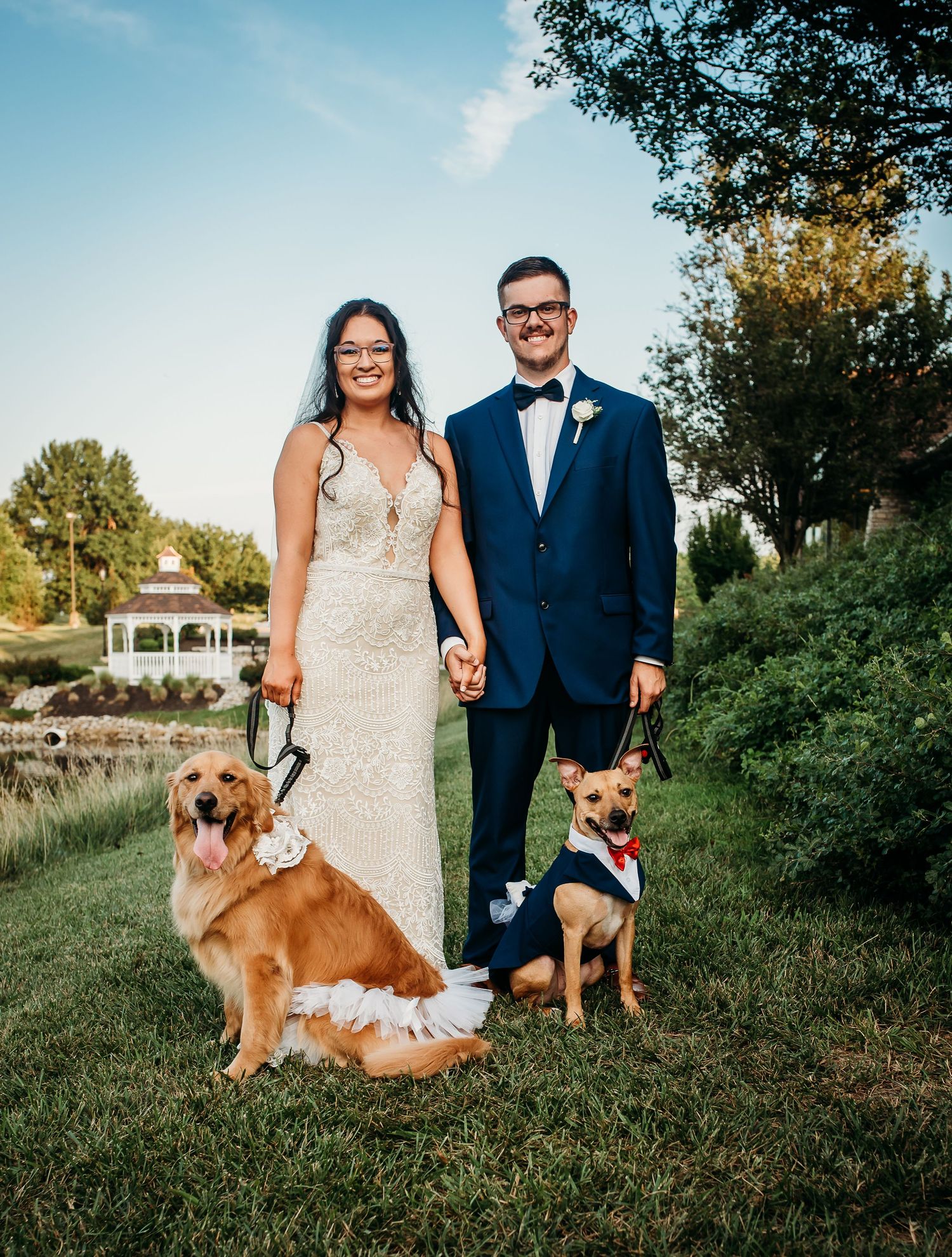 A newlywed couple poses with their two dogs in formal wedding attire outdoors.
