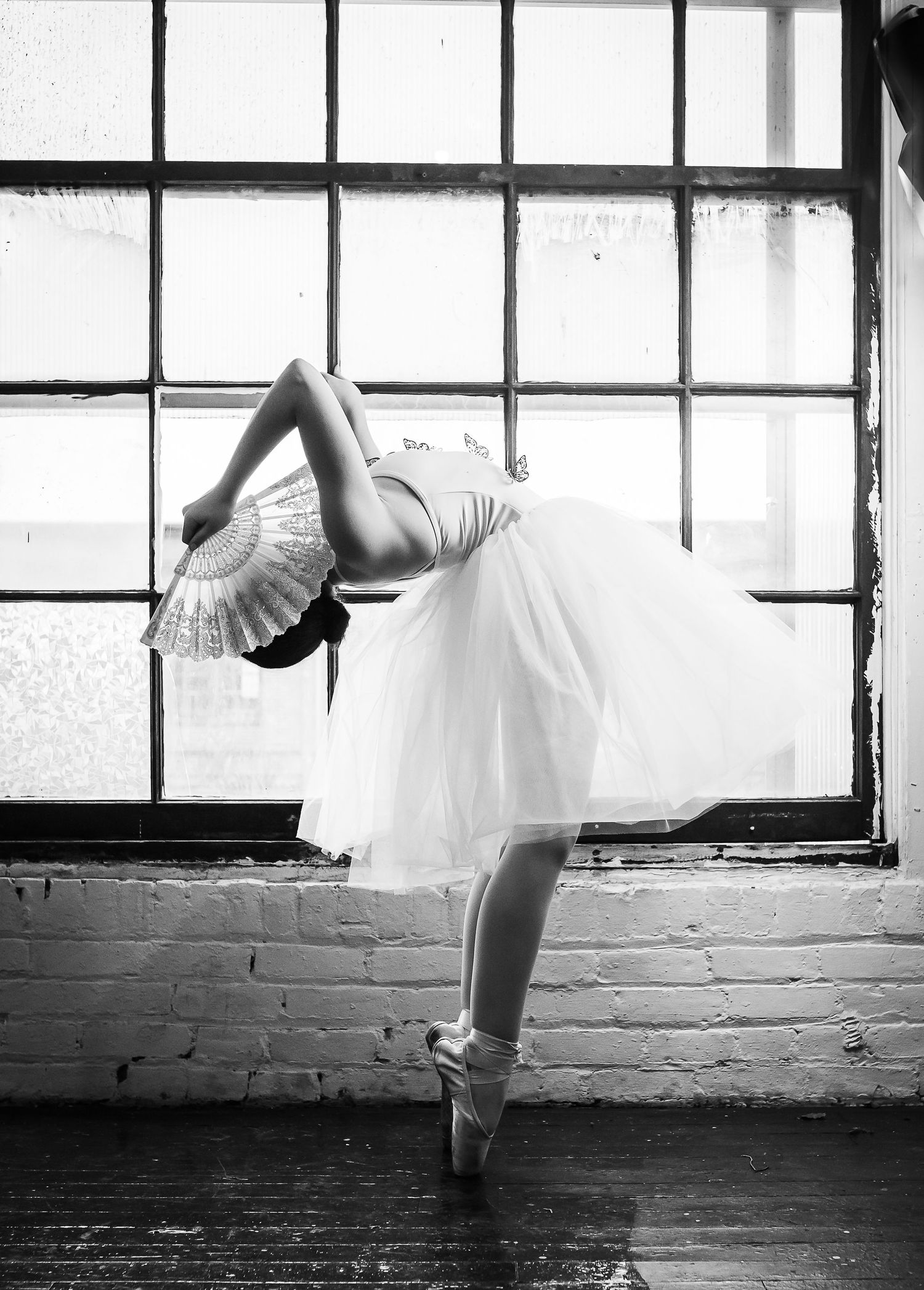 A ballerina in a white tutu performs a graceful dance pose against a window backdrop in black and white.