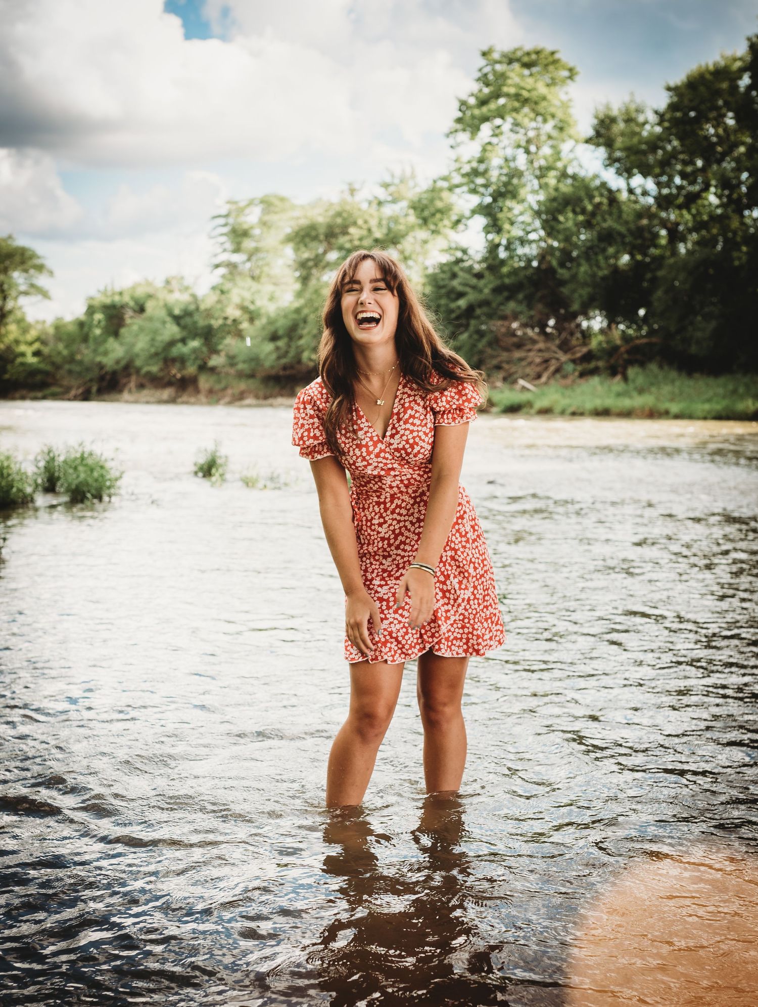 Woman in floral dress playing in shallow river water with cloudy sky backdrop.
