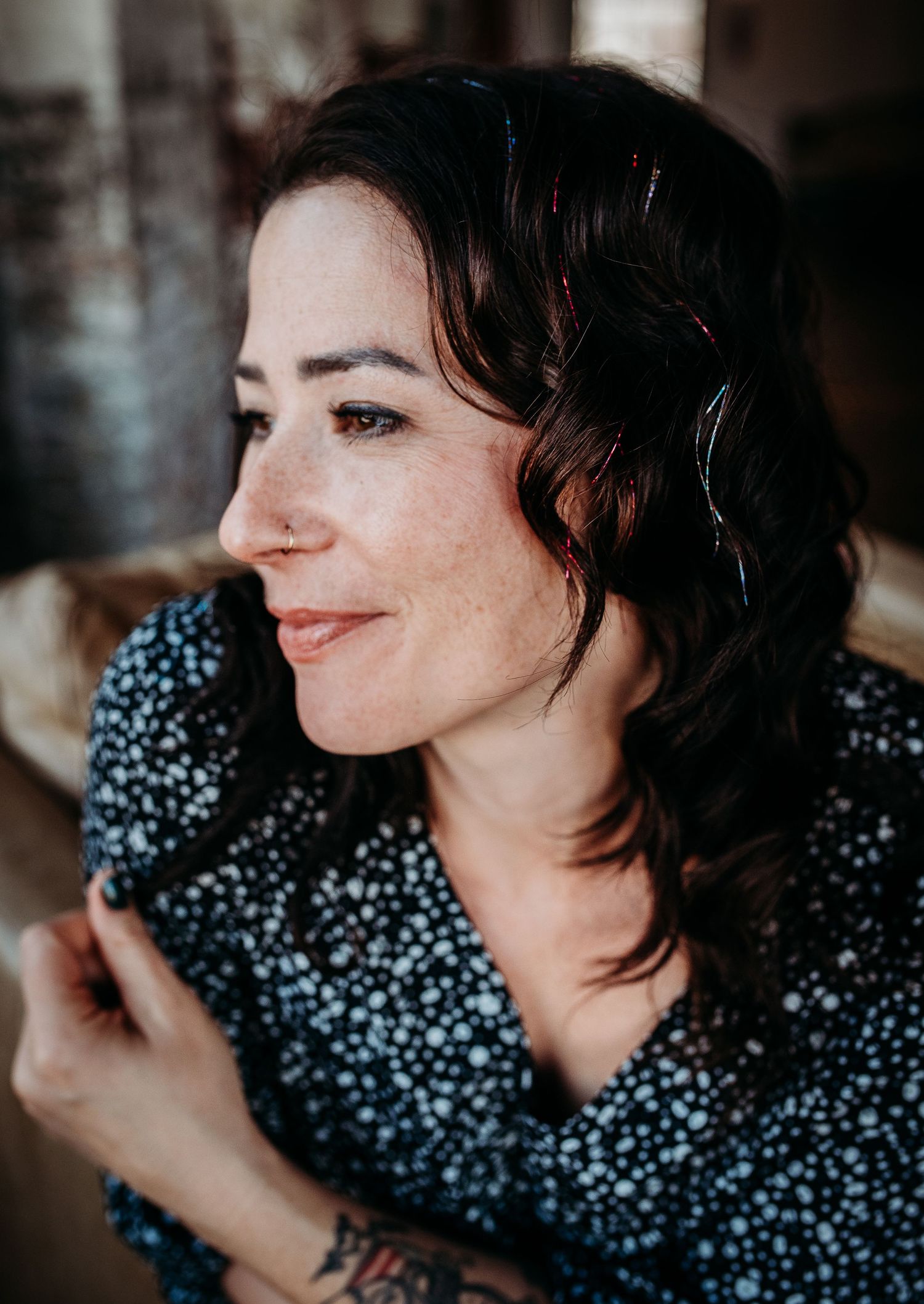A woman in a polka dot dress smiles while looking off to the side in soft lighting.