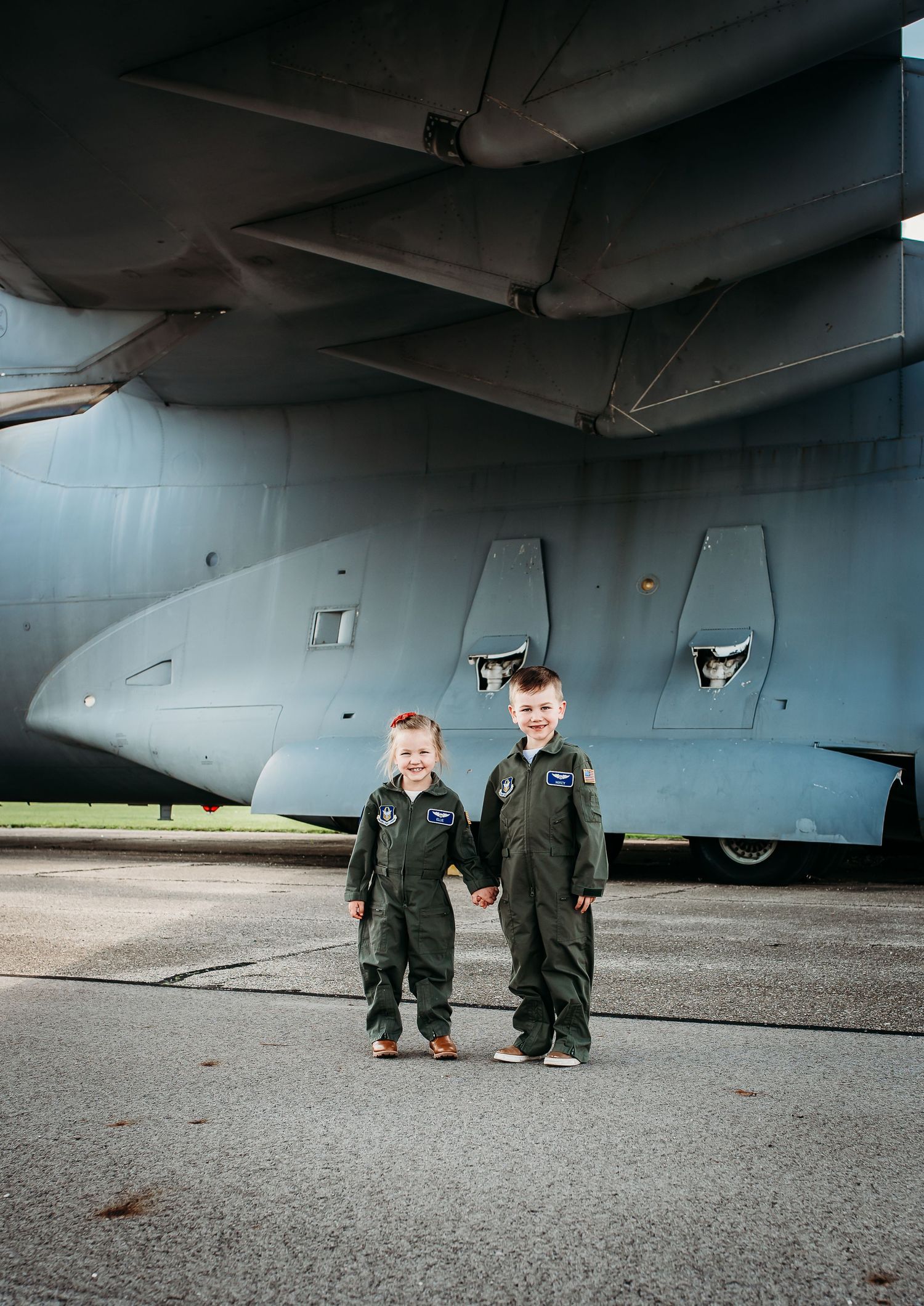 Two children wearing oversized military uniforms stand beneath a large military aircraft wing.
