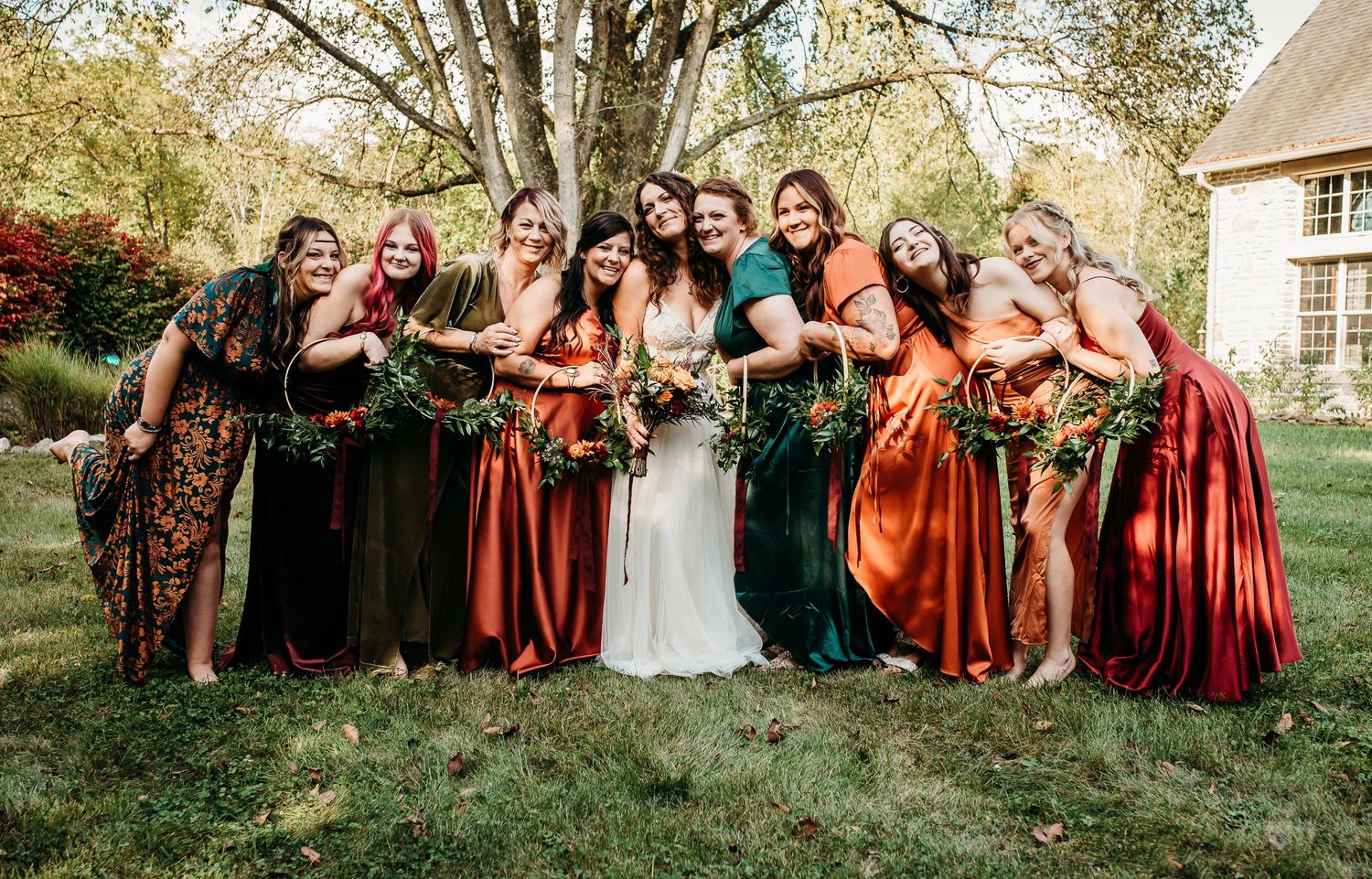 Wedding group photo with bridesmaids in autumn-colored dresses gathered under trees.