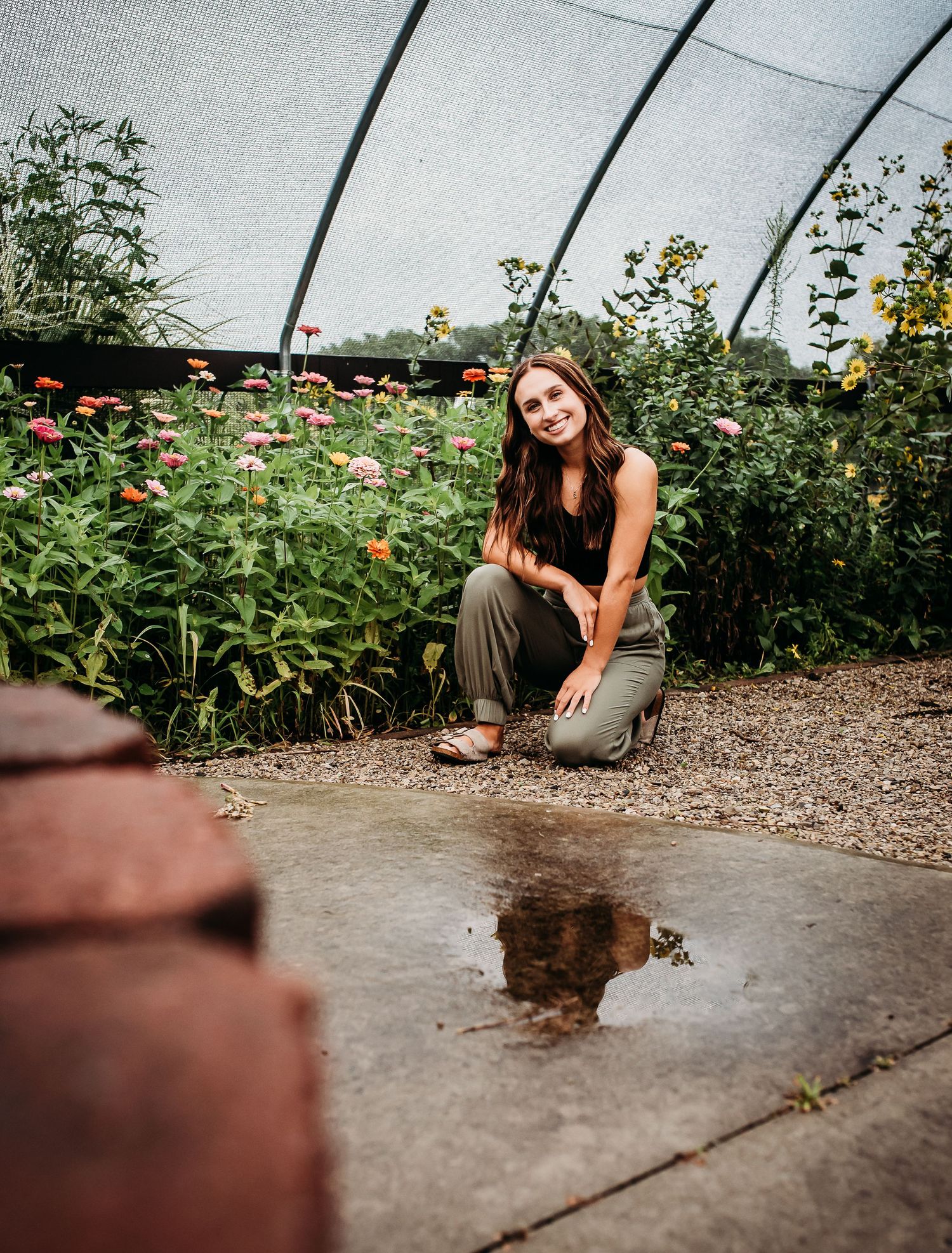A person in black sits by a garden bed with flowers and muddy puddle reflections.
