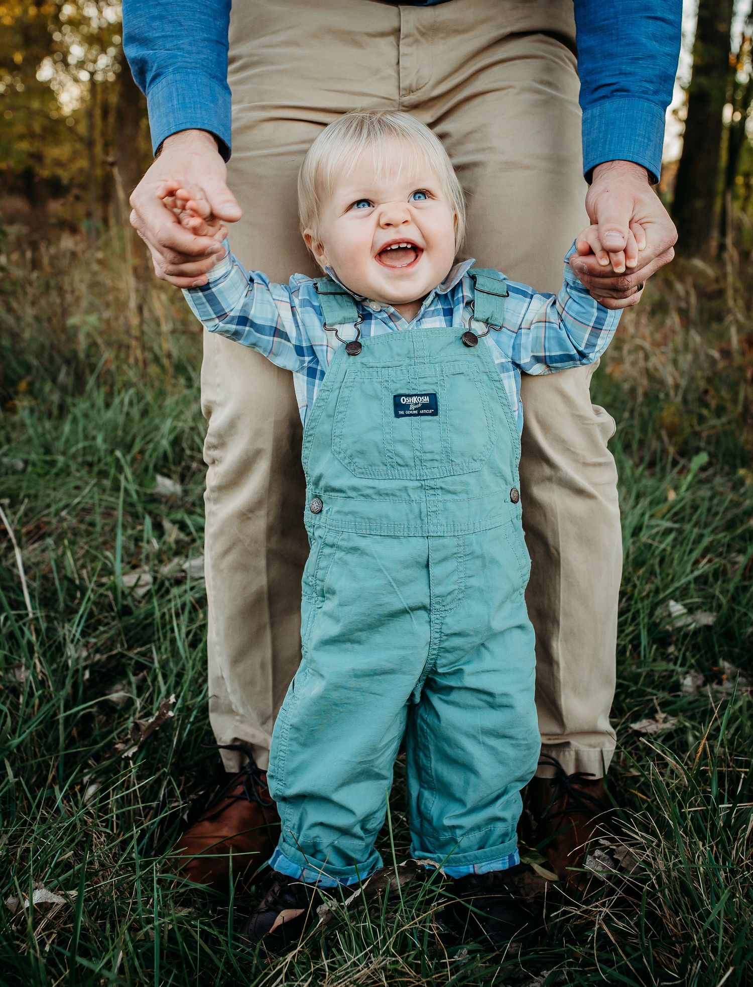 A laughing toddler in teal overalls holding hands with someone on grass.