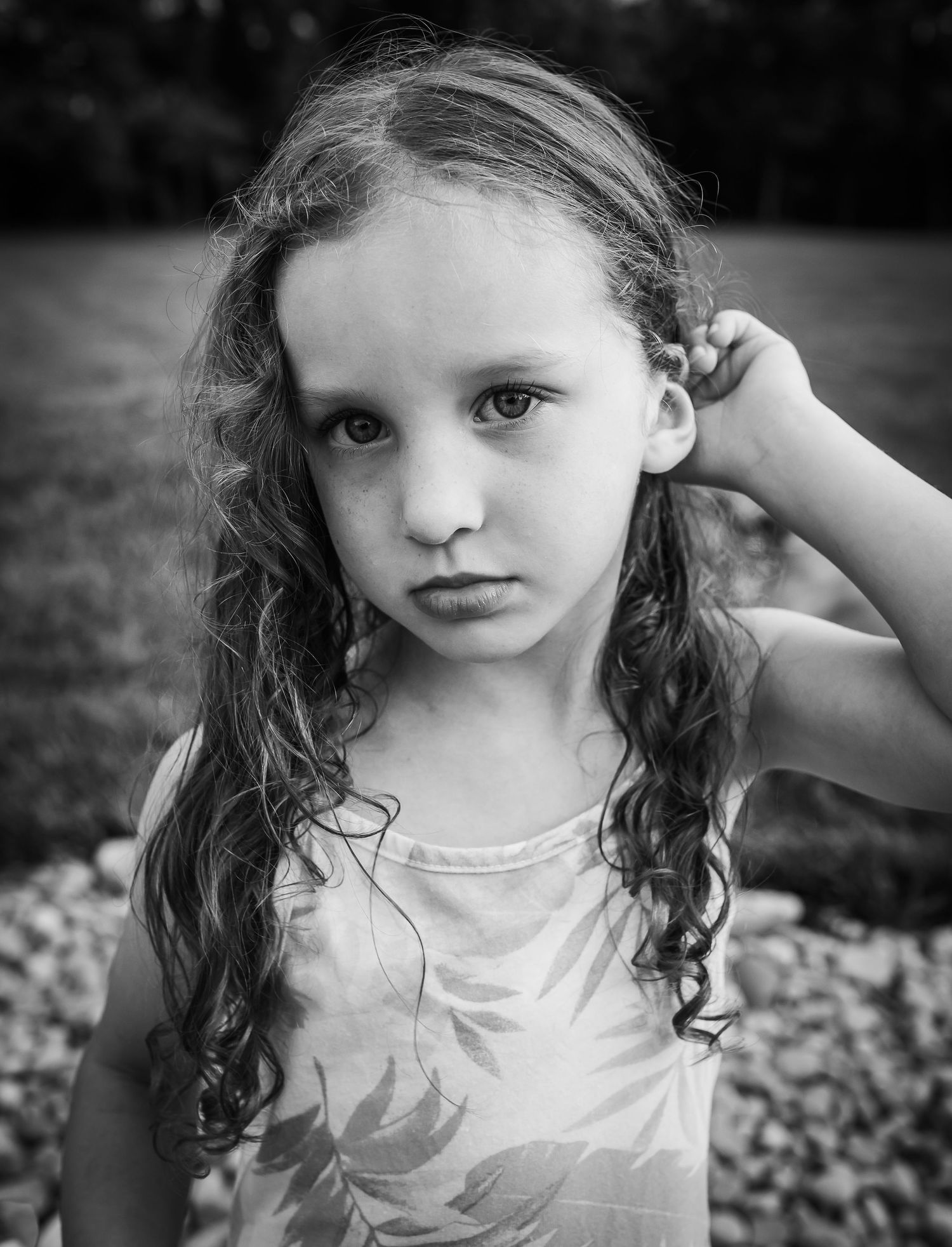 Black and white portrait of a child with long curly hair and serious expression wearing a light colored top outdoors.