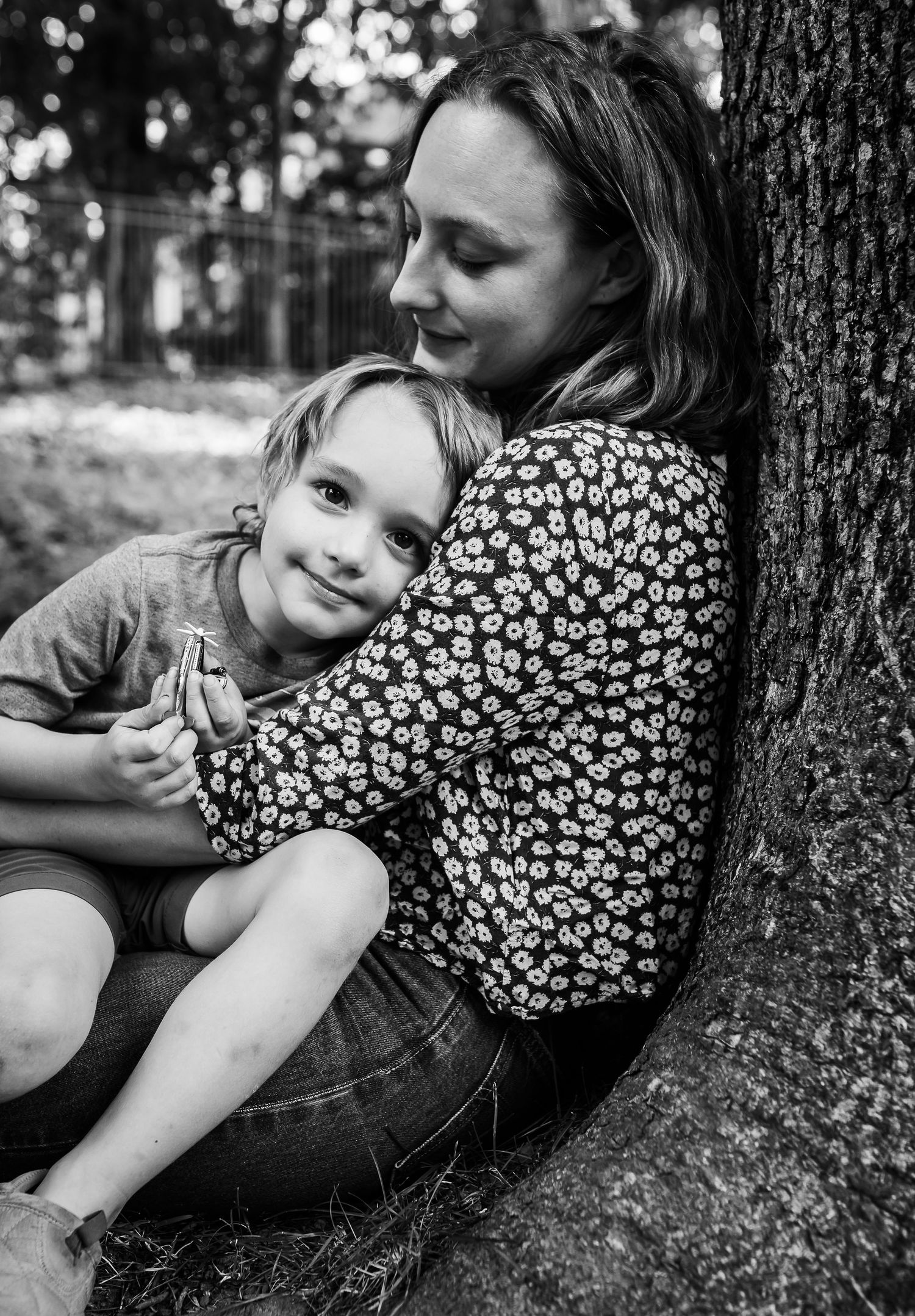 A tender moment between parent and child as they rest against a tree trunk in a black and white photograph.