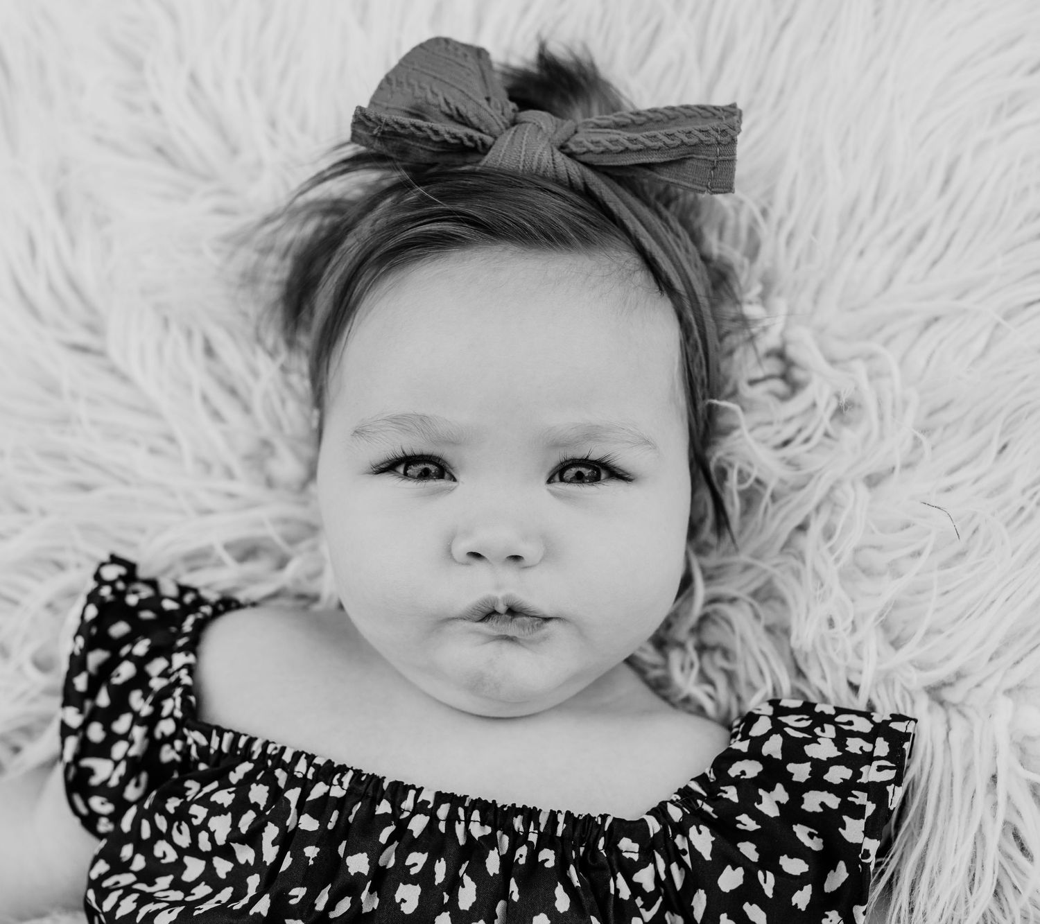 A baby wearing a polka dot dress and bow headband lies on white fluffy fabric.