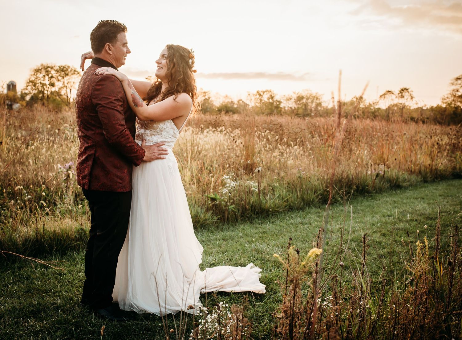 A romantic wedding couple embraces in a tall grass field during sunset.