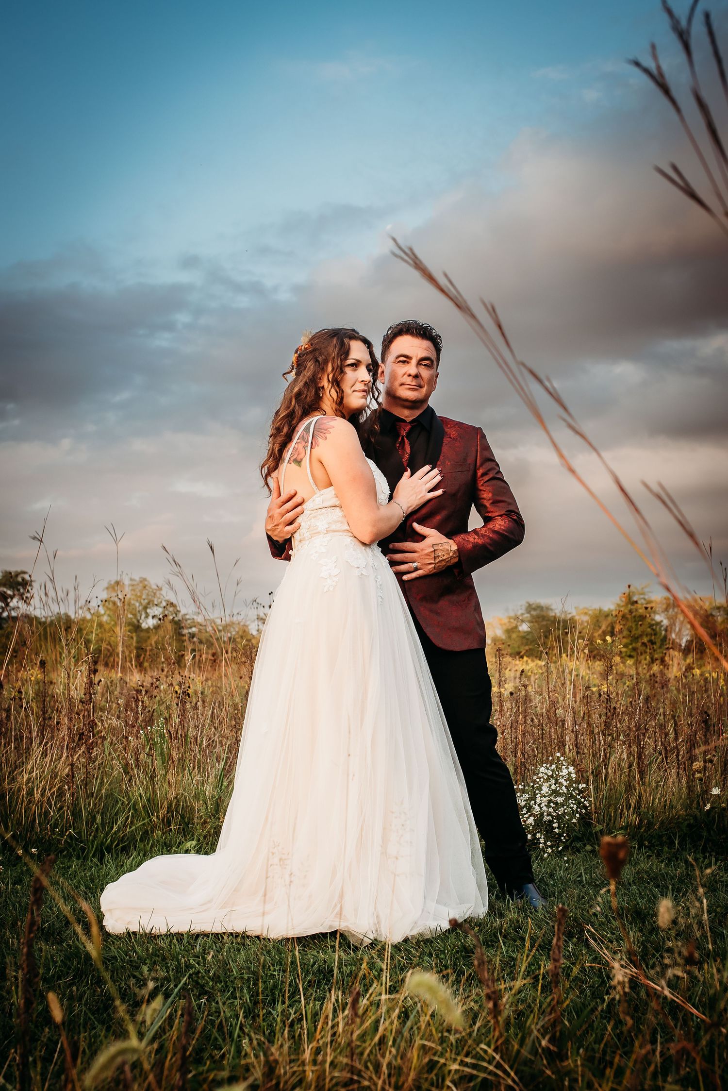 A romantic wedding couple embraces in a field at sunset with tall grass and moody sky.