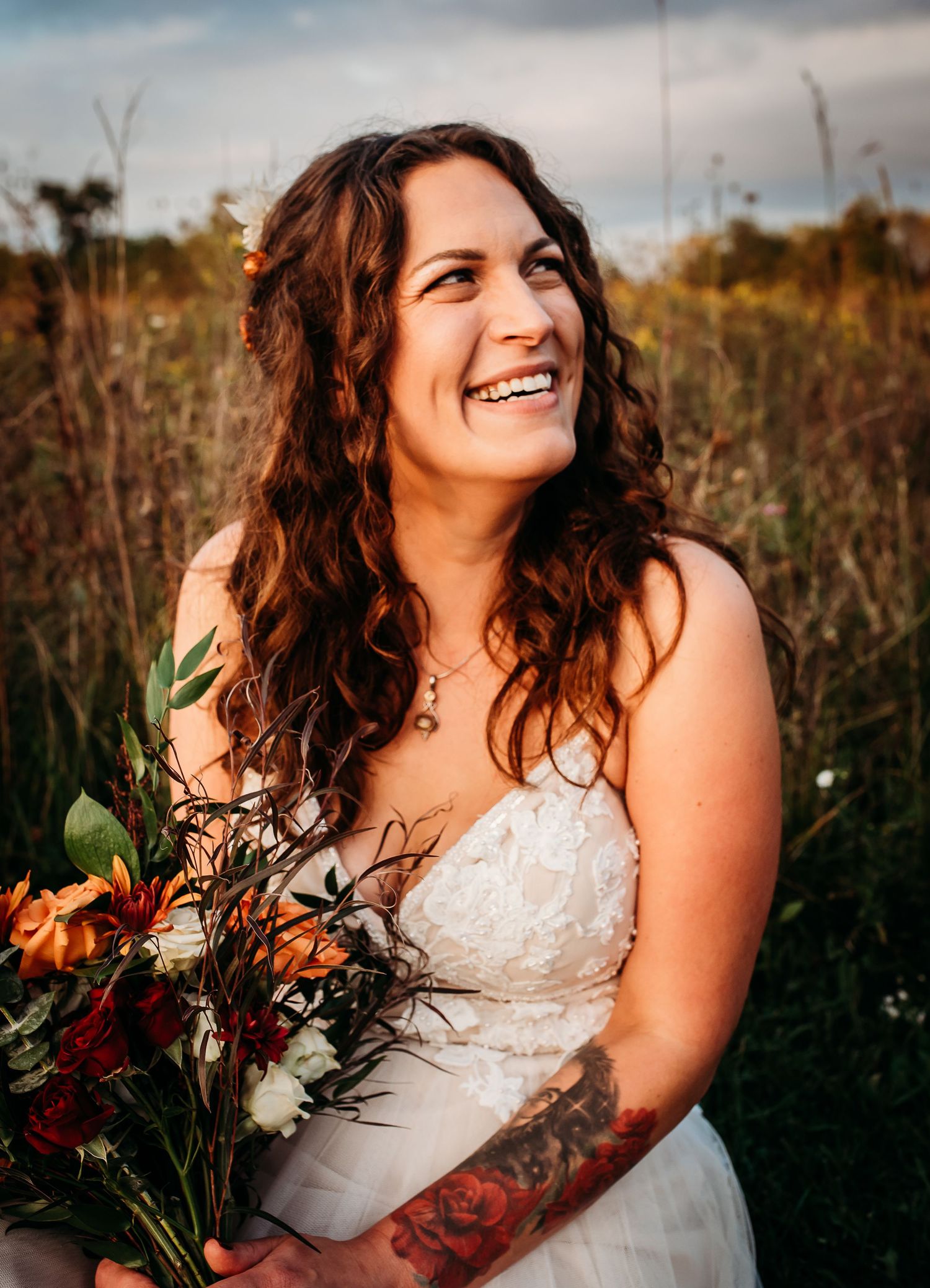 A bride in white dress holds red flowers while laughing in a rustic outdoor setting.