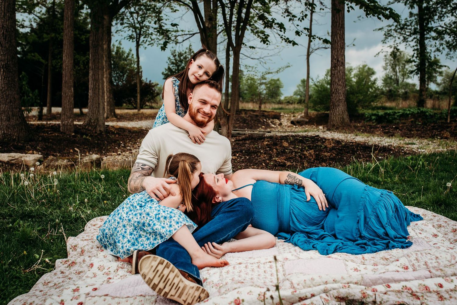 A family relaxes together on a patterned blanket in a wooded setting.