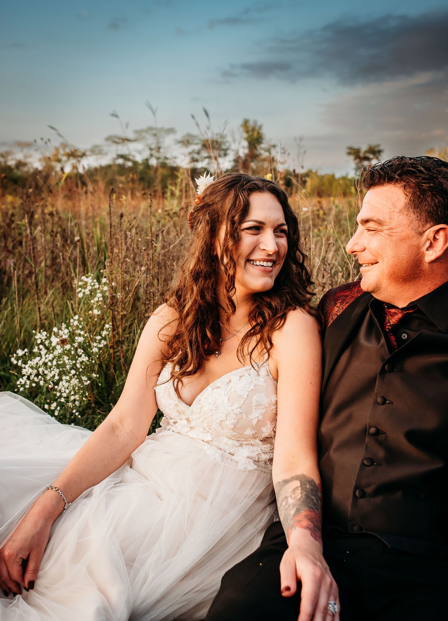 A joyful couple in wedding attire sits together in a golden field at sunset.