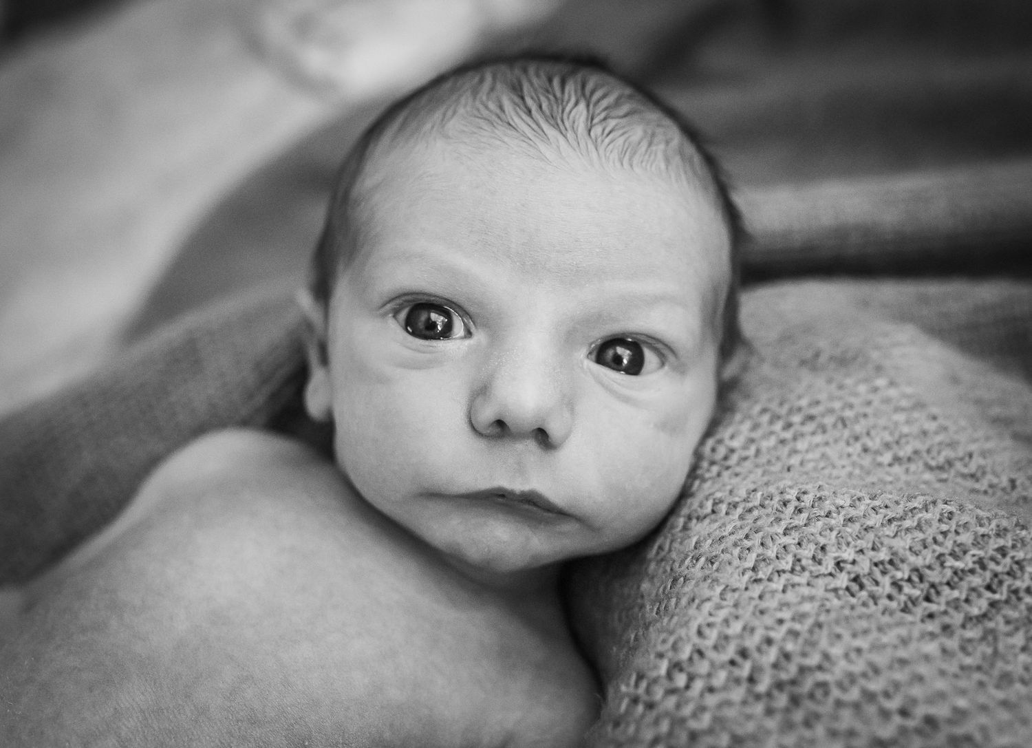 Close-up black and white portrait of a newborn baby with wide-eyed expression.