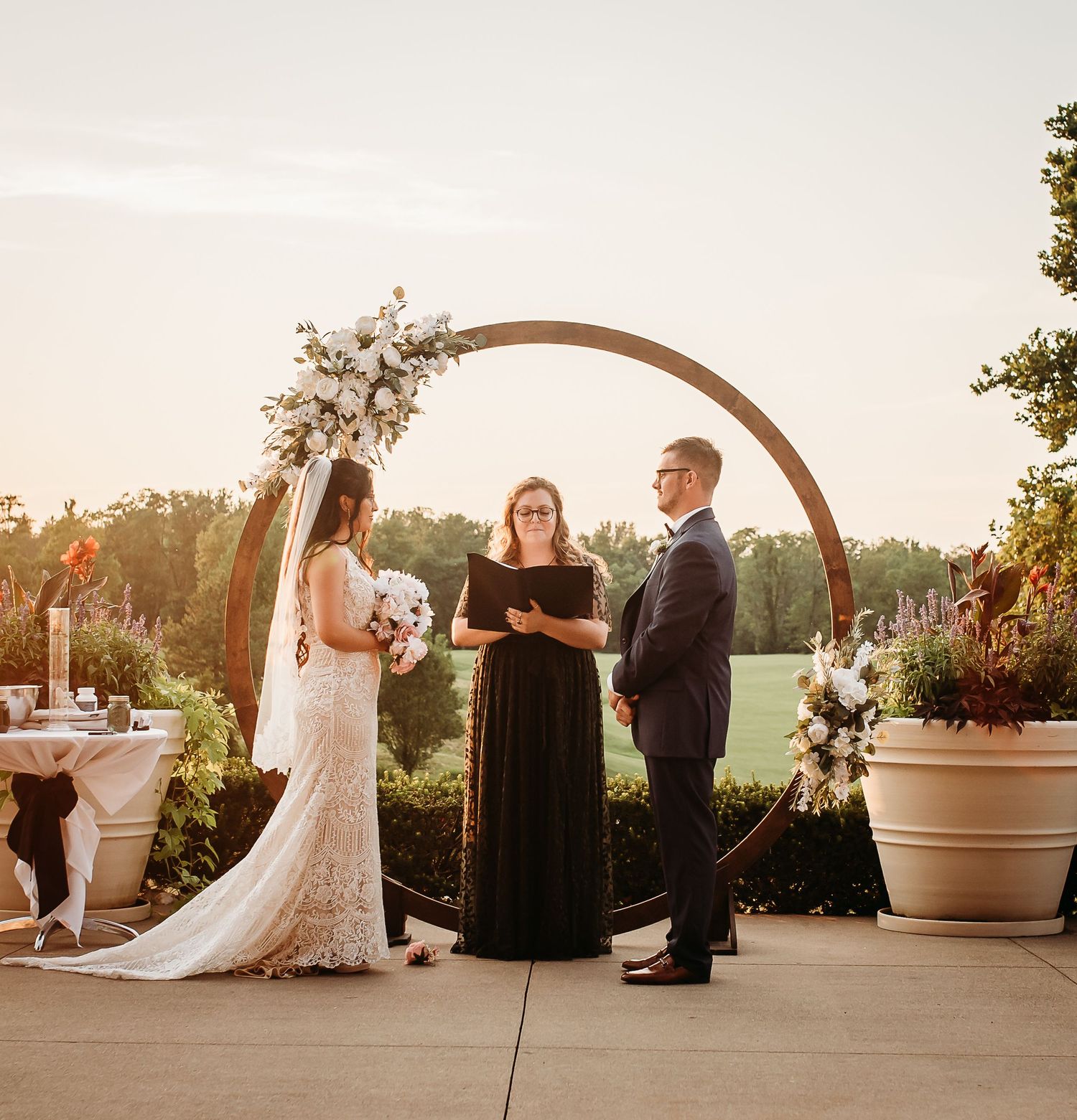 A wedding ceremony under an arch decorated with white flowers at sunset.