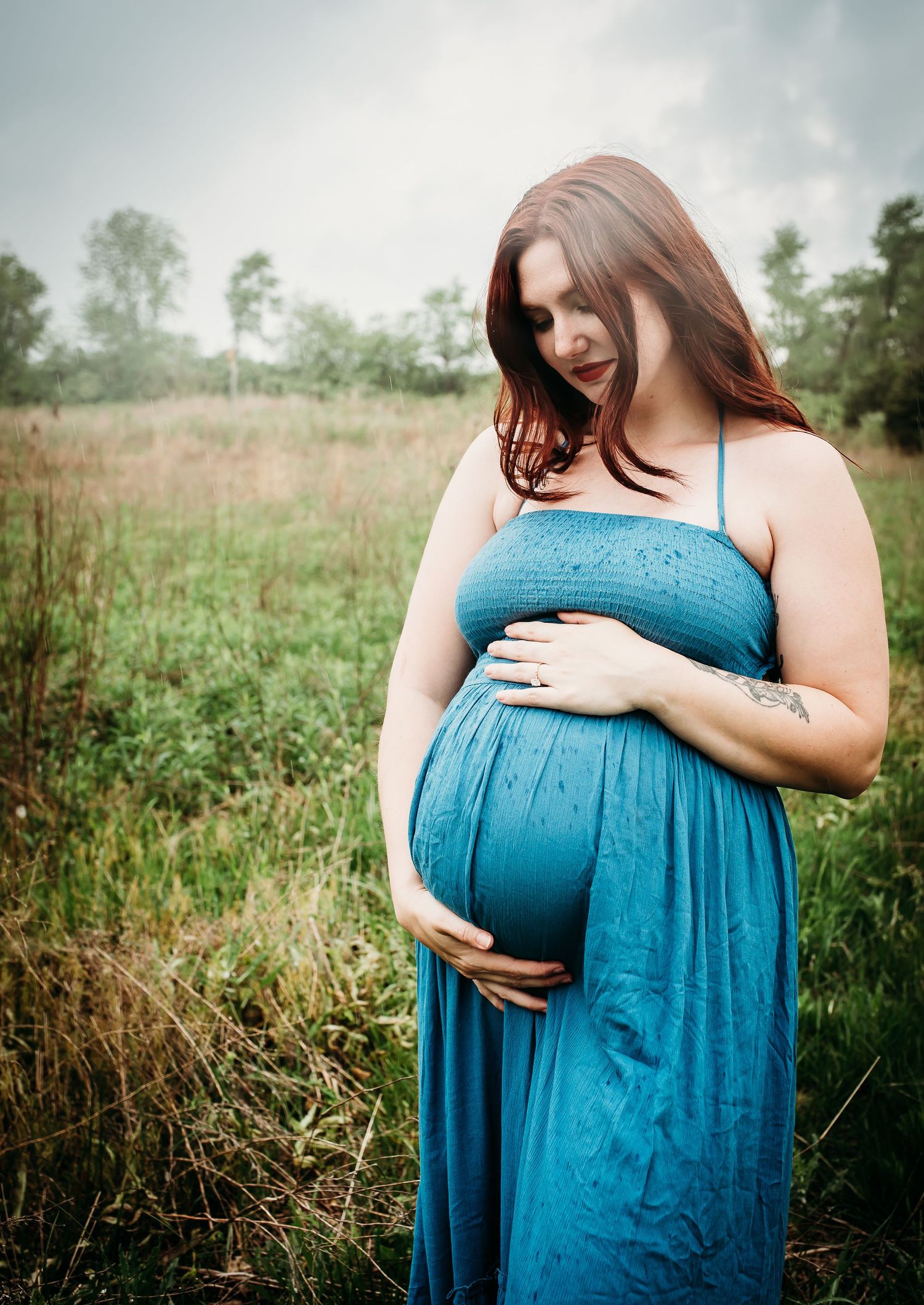 A pregnant woman in a flowing blue dress cradles her belly in a natural outdoor setting.