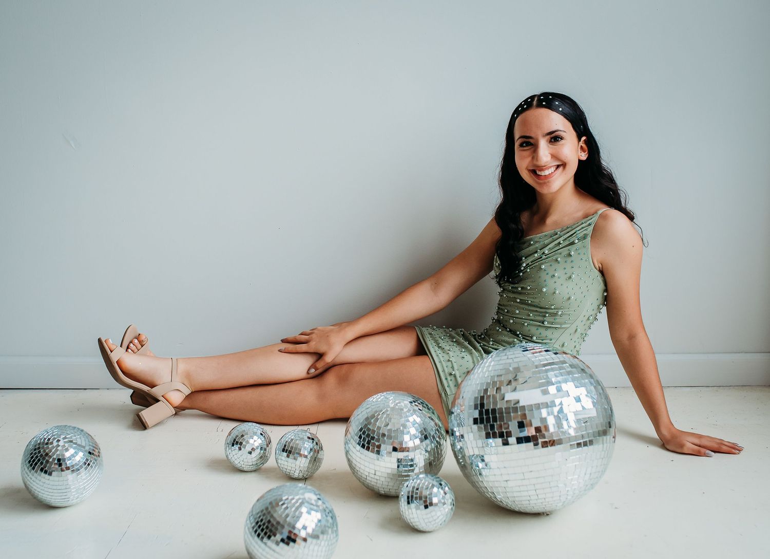 A festive scene with woman in green dress posing with multiple disco balls on light background.