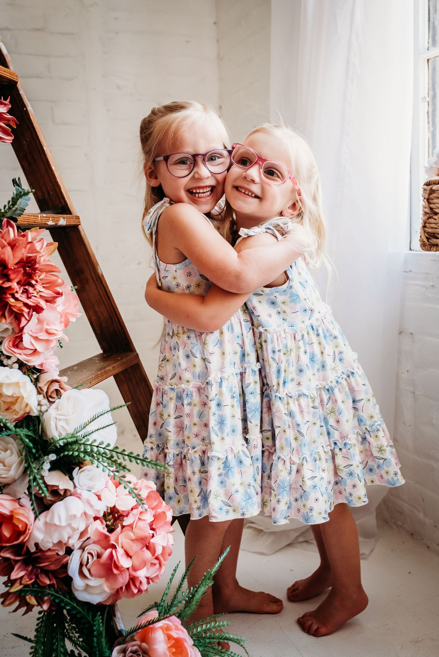 Two young children in matching floral dresses embrace and laugh in a bright white studio.