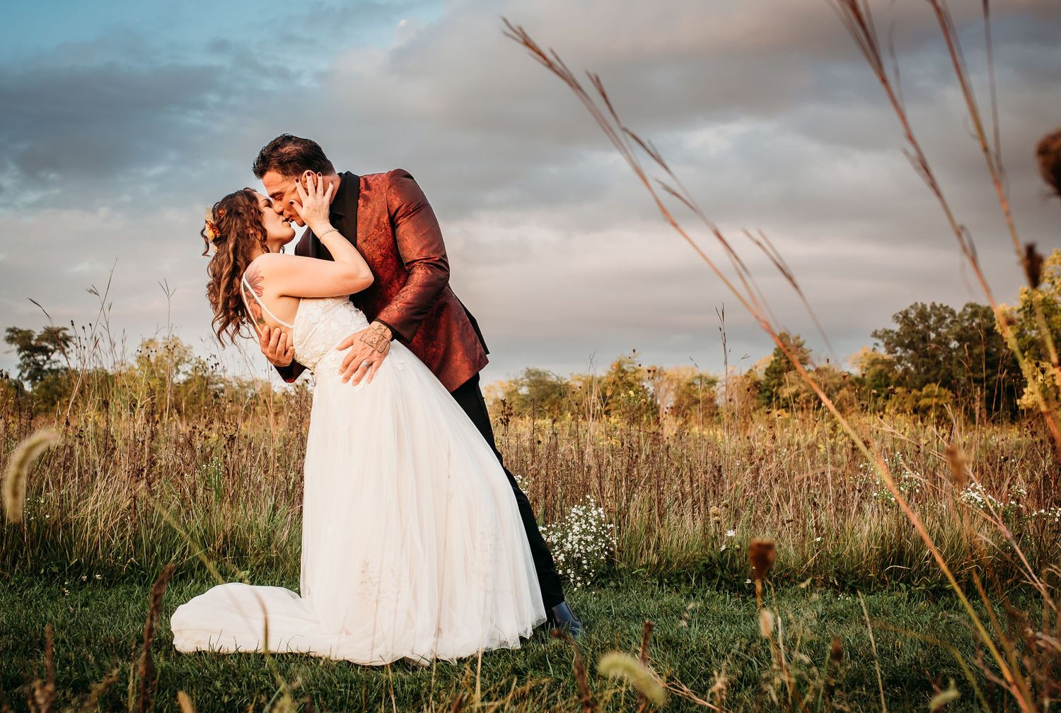 A romantic wedding couple dips in dramatic pose among tall grass at sunset.