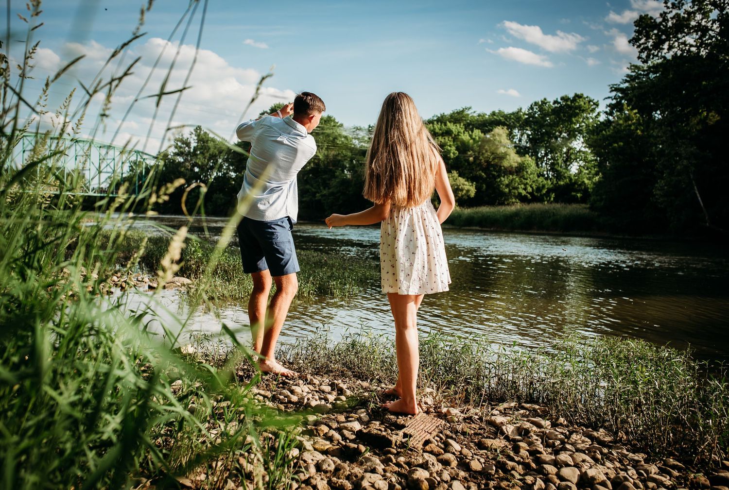 Couple standing on rocky riverbank surrounded by tall grass on sunny day.