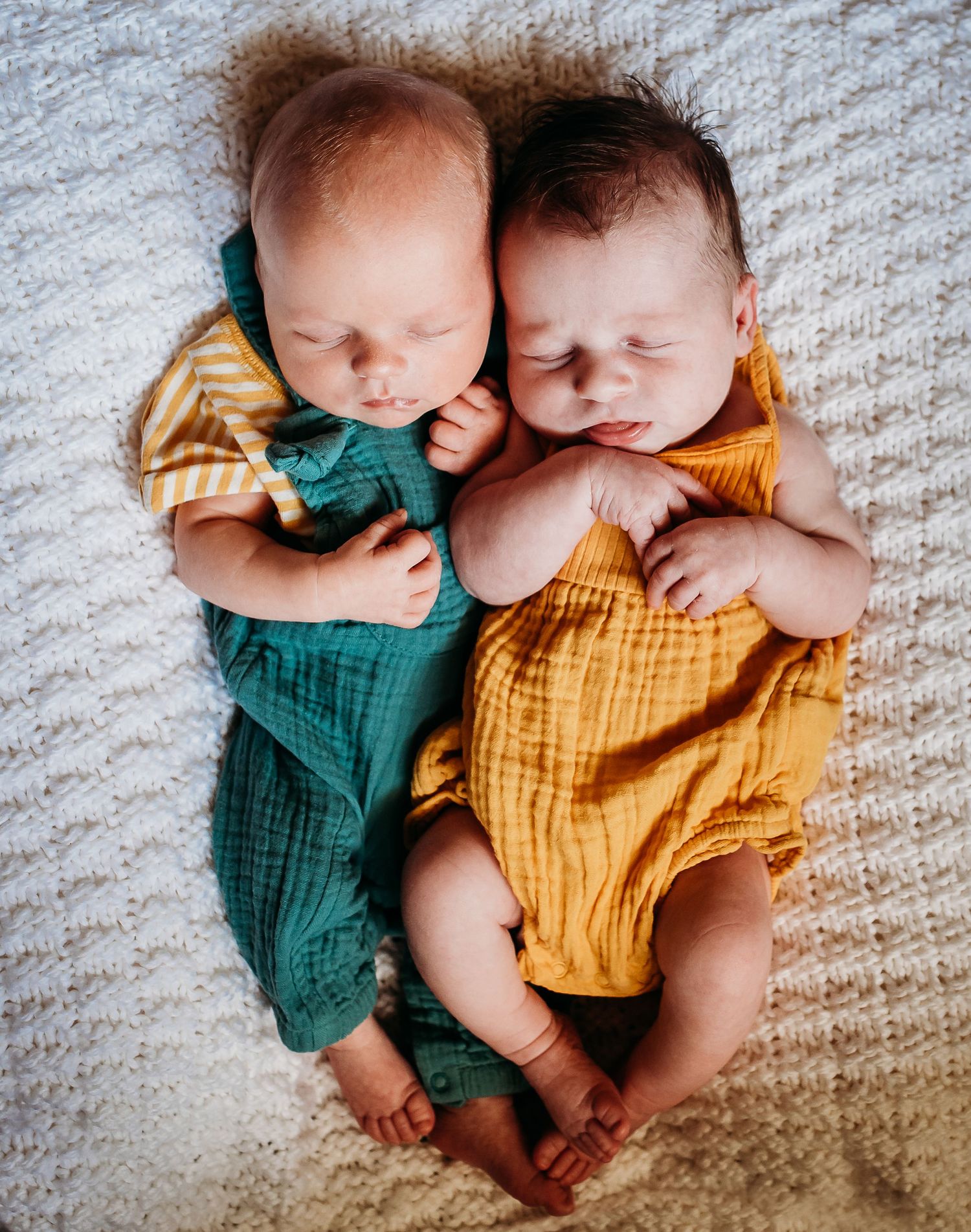 Two sleeping newborn babies wearing green and yellow rompers lying on textured blanket.