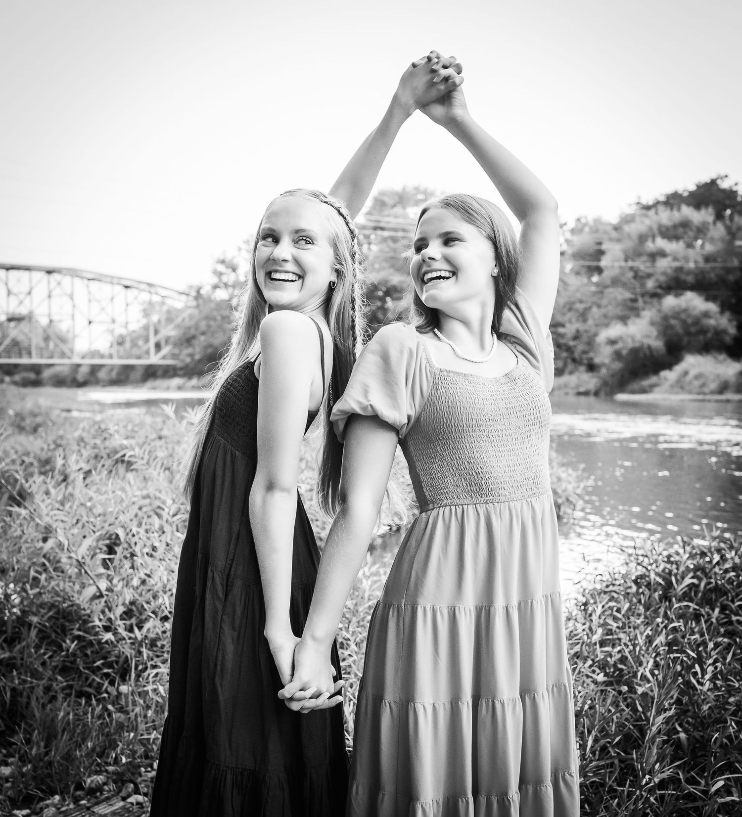 Two friends in flowing dresses dance and laugh together by a river in black and white.