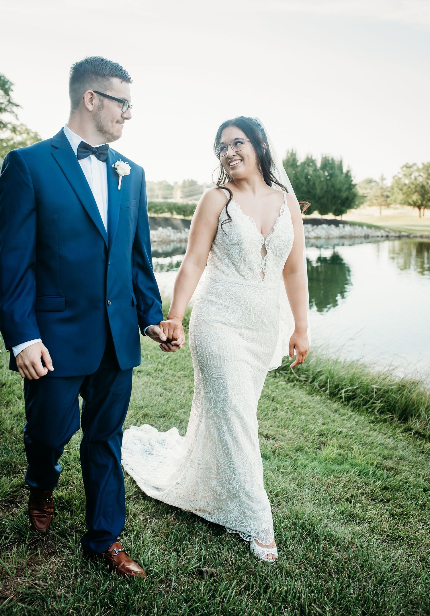 A wedding couple walks hand in hand along a peaceful waterfront path.