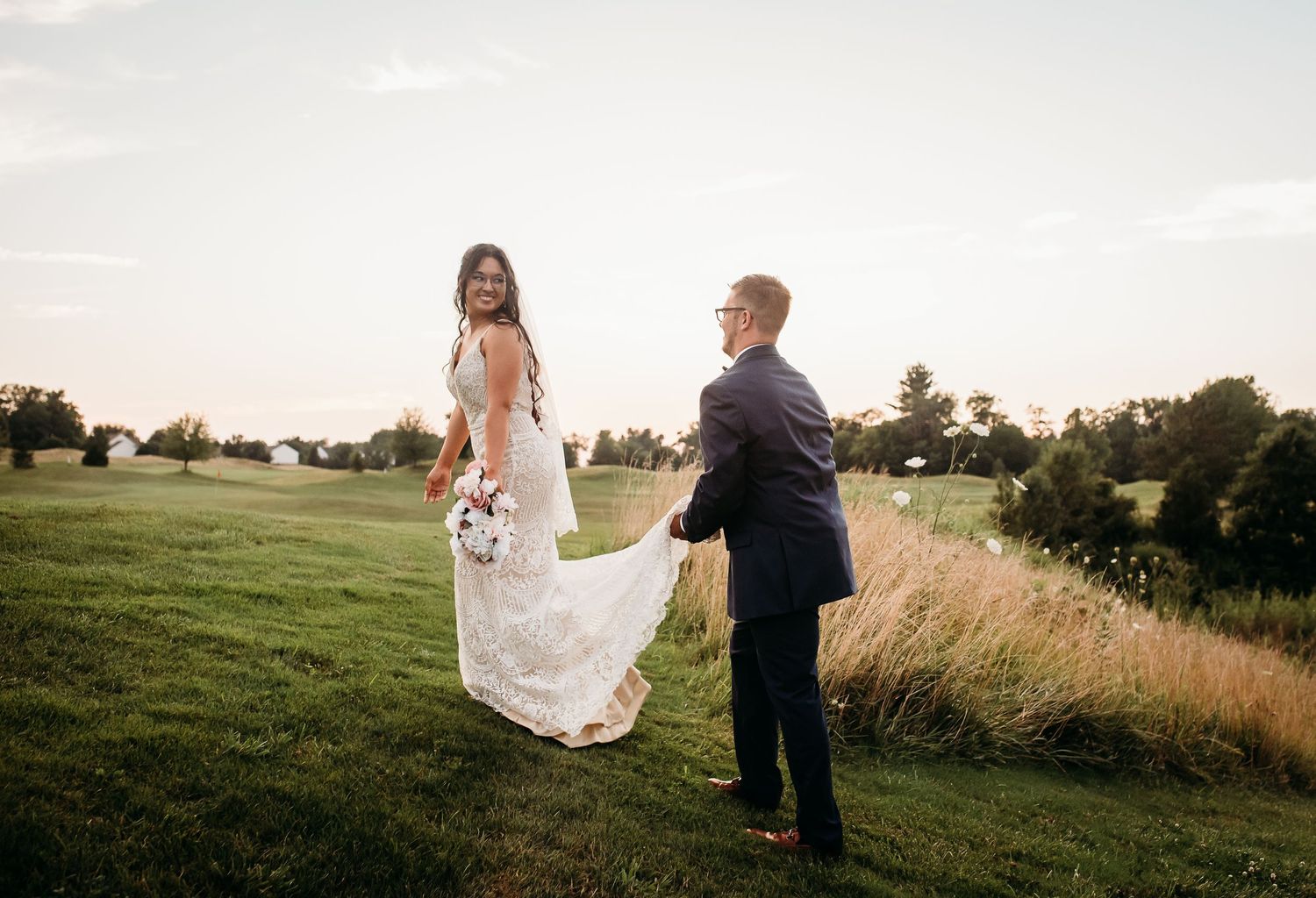 Couple in wedding attire walking together through golden grass at sunset in an outdoor field setting.