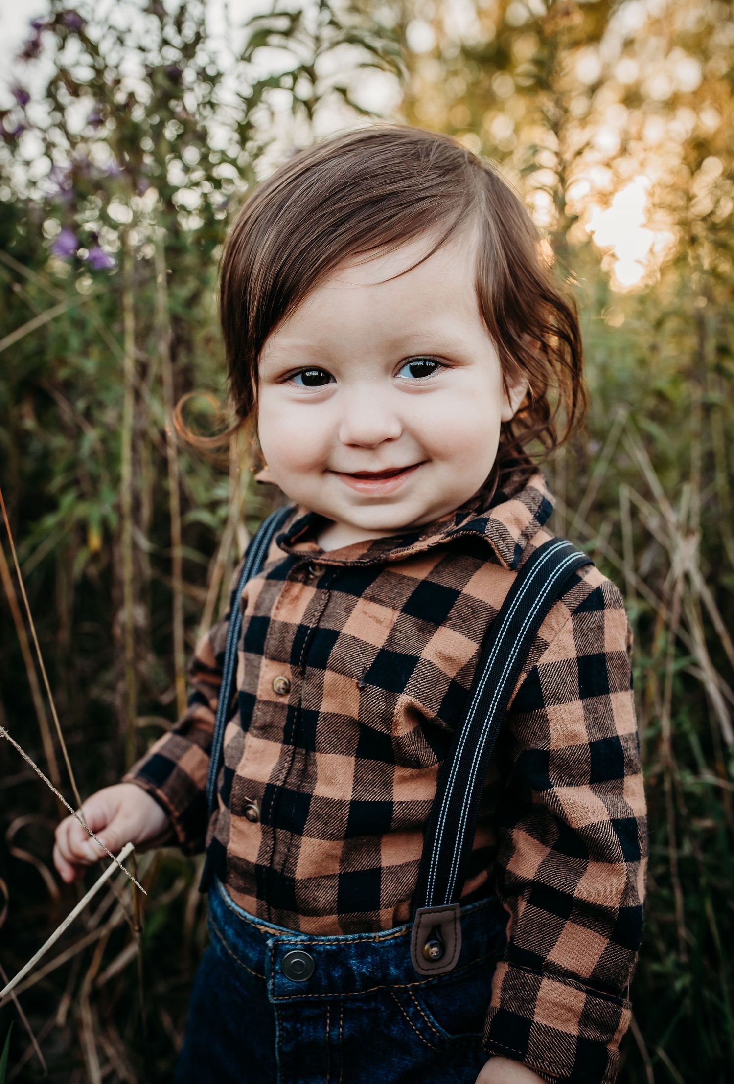 Small child wearing plaid shirt and overalls smiling outdoors at sunset.