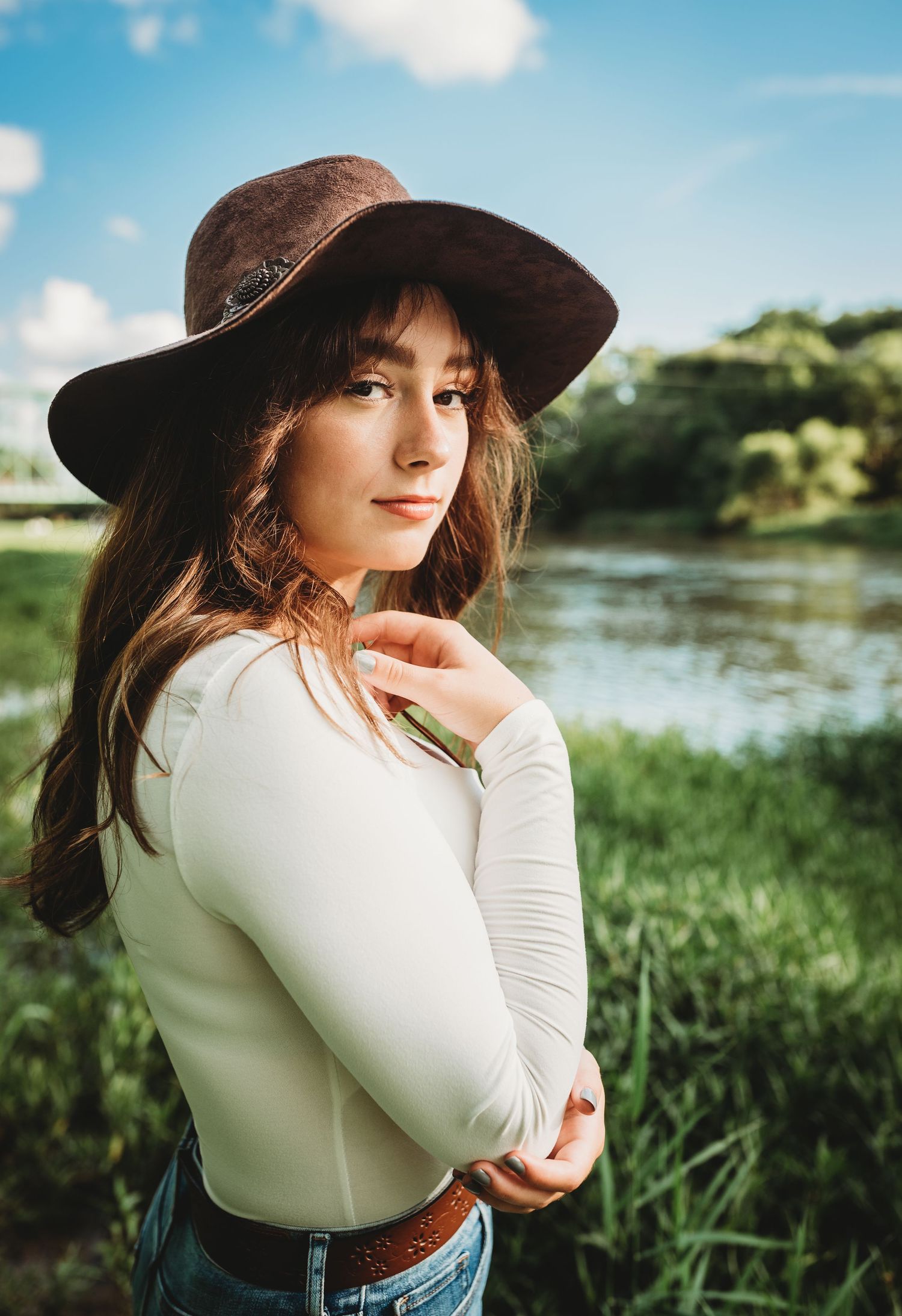 Person in white top and wide-brim hat poses thoughtfully by riverside with lush greenery.