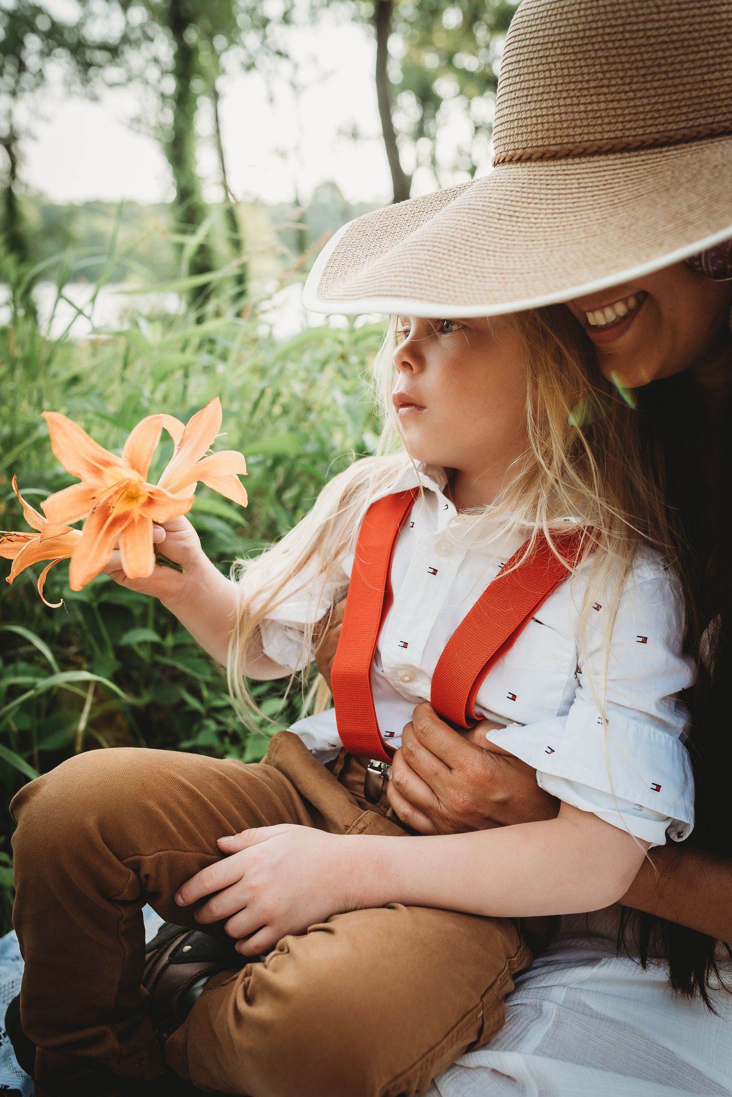 Child holding orange lily flower while sitting outdoors in sunhat.