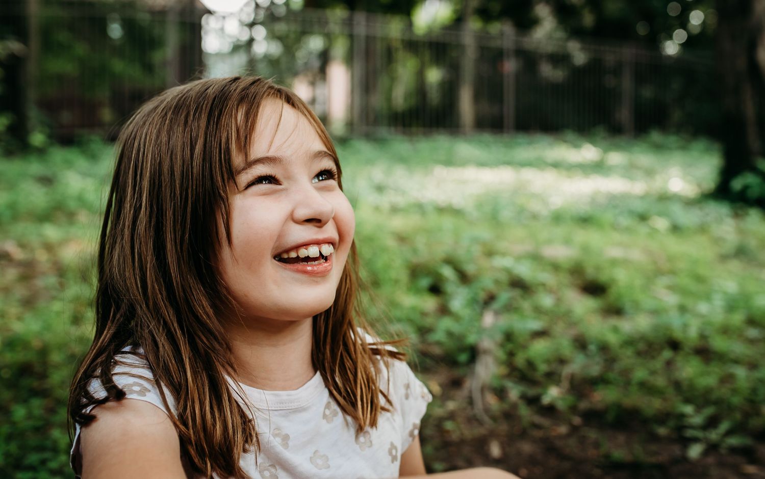 A person laughs joyfully while outdoors on a sunny day with greenery in the background.