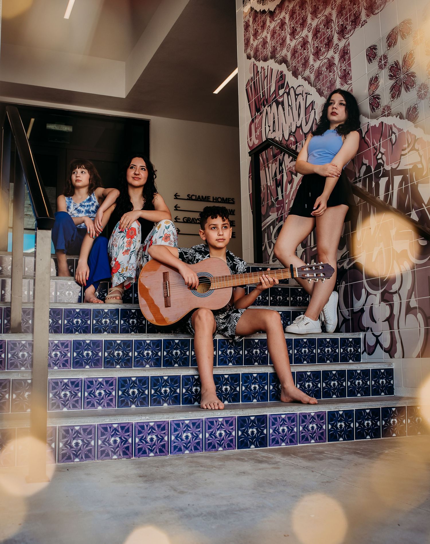 A group of musicians pose on decorative tiled steps with a guitar while wearing casual summer outfits.