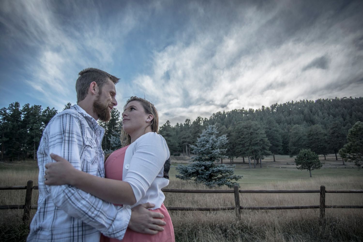 A couple sharing an intimate moment against a stormy sky backdrop with rustic fence in background.